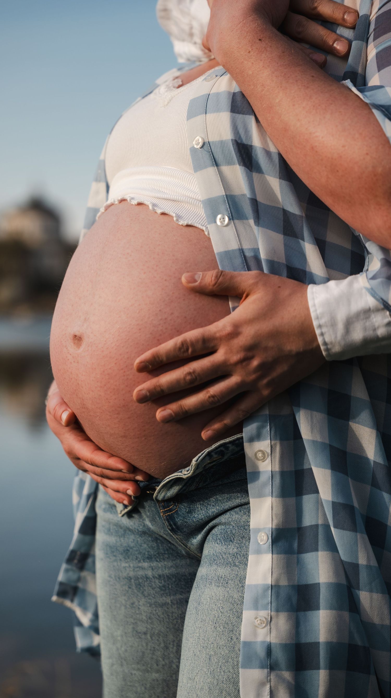 Couple tenderly cradling pregnant belly outdoors near water, woman in plaid shirt and jeans.