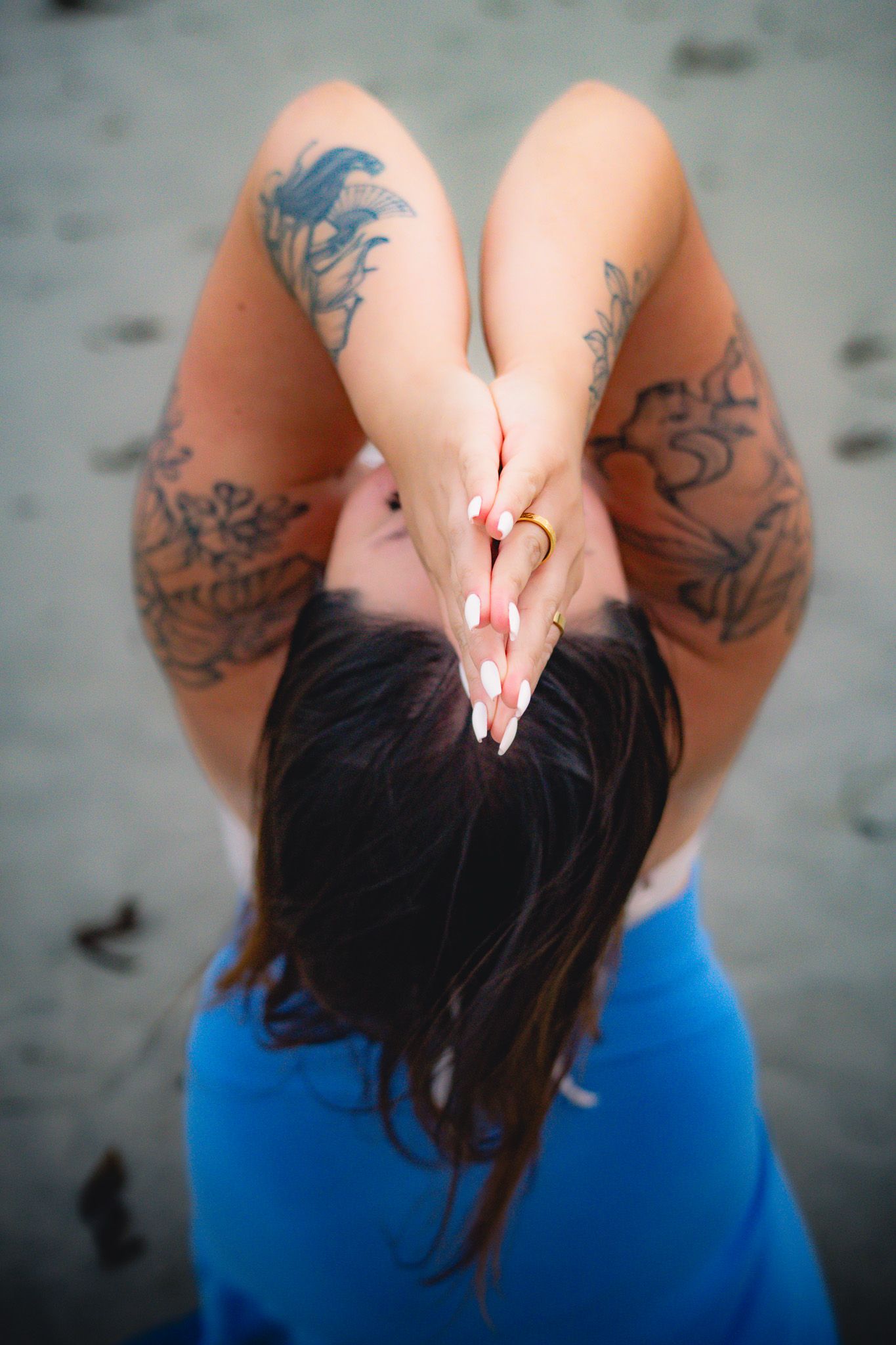 Woman with tattoos on arms clasps hands behind head, wearing blue top and gold ring, viewed from above.