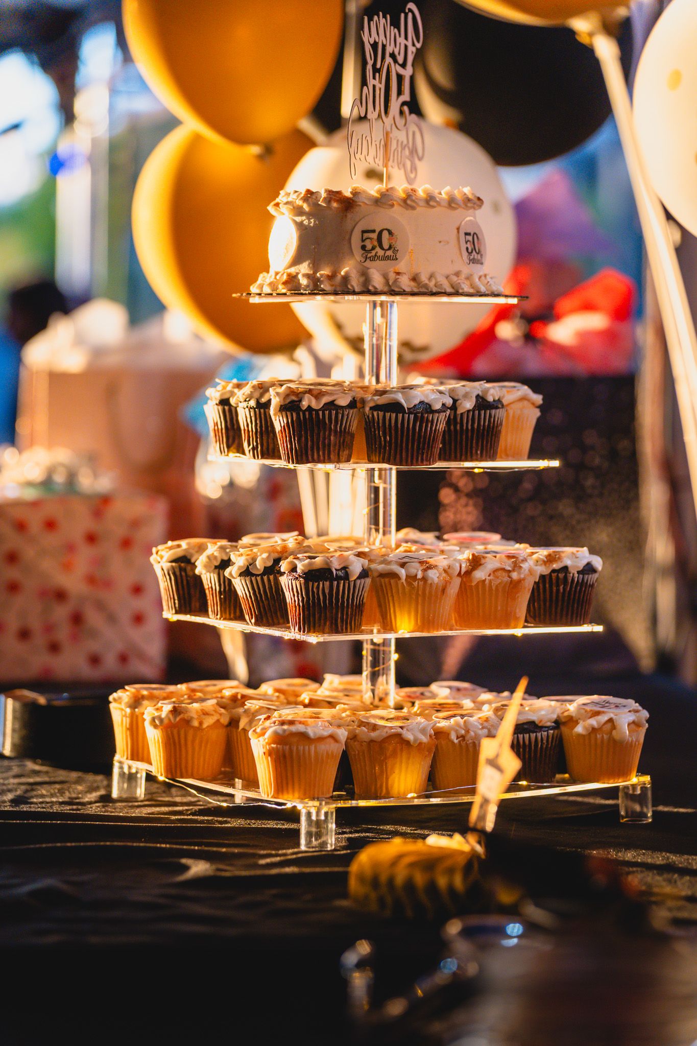 Tiered cupcake tower display with a small birthday cake on top, surrounded by gold and black balloons at a celebration.