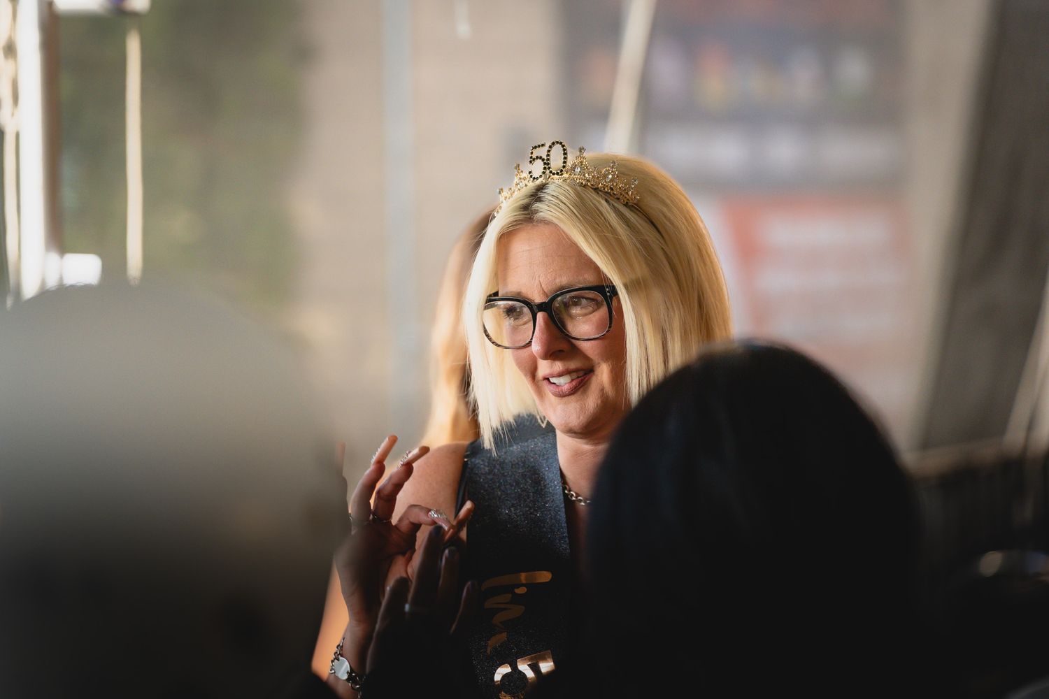 Woman wearing 50th birthday tiara and glasses smiling at a celebration party.