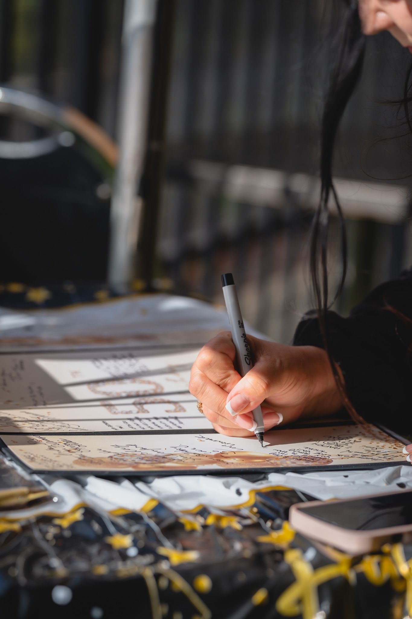 Close-up of a person writing on illustrated papers with a pencil at an outdoor table with scattered ribbons.
