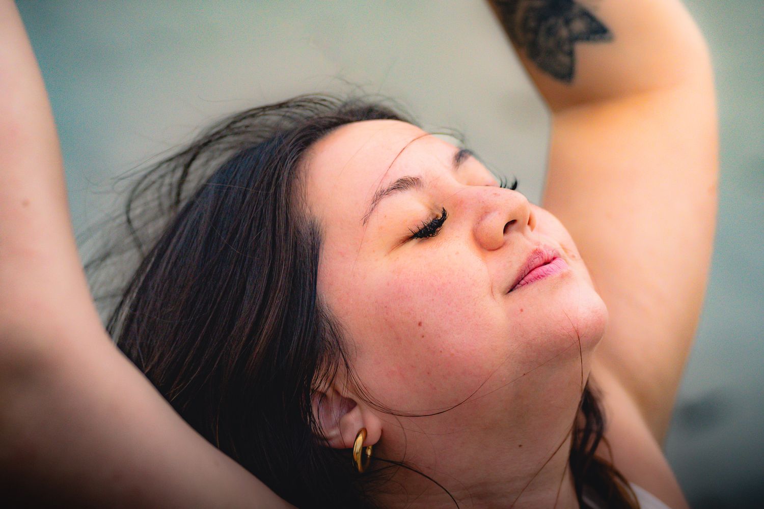 Close-up of a woman with dark hair, closed eyes, and gold hoop earrings, with a tattoo visible on her arm.