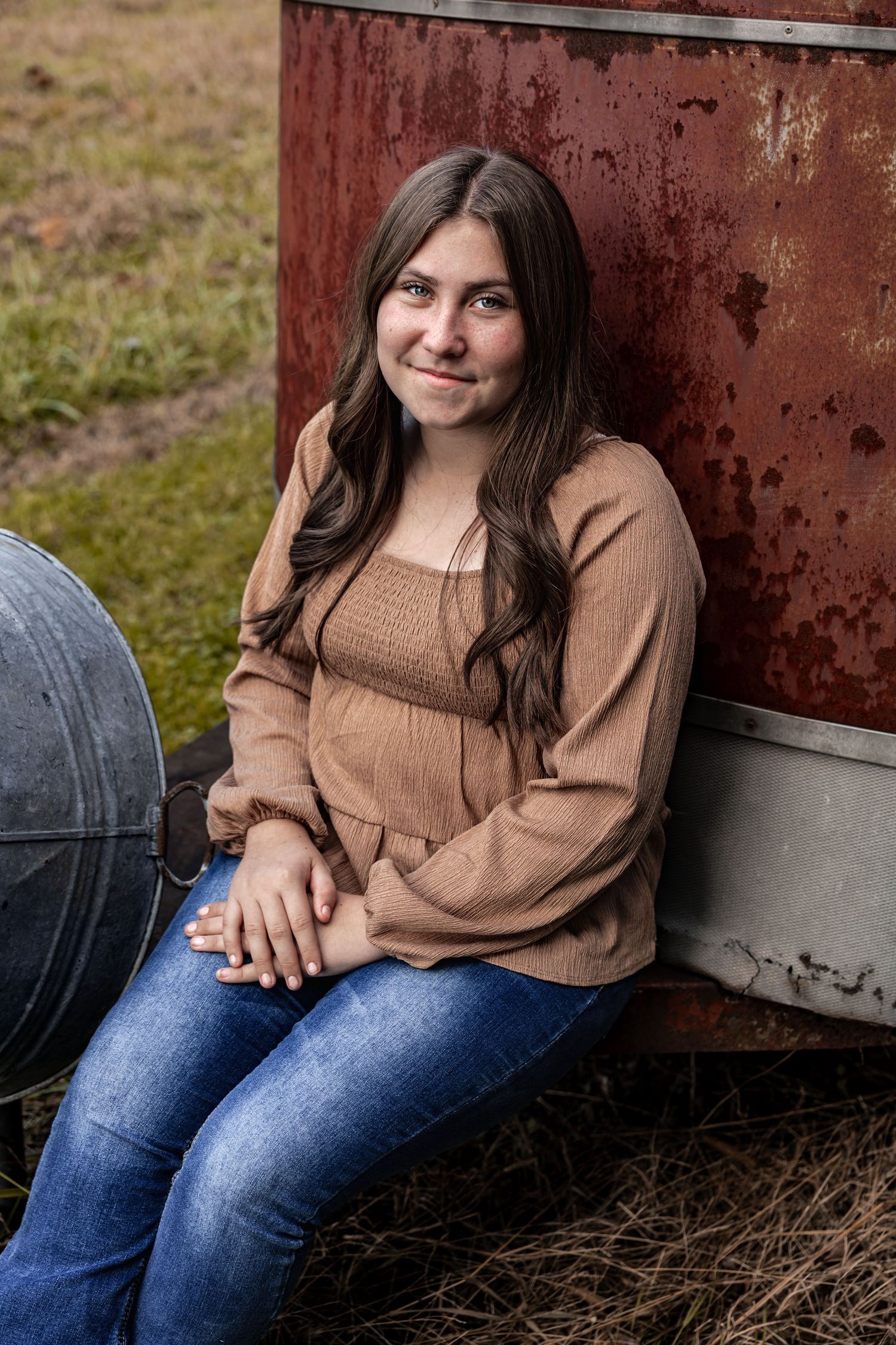 A person in a brown sweater and jeans sits casually against a rustic red metal structure outdoors.