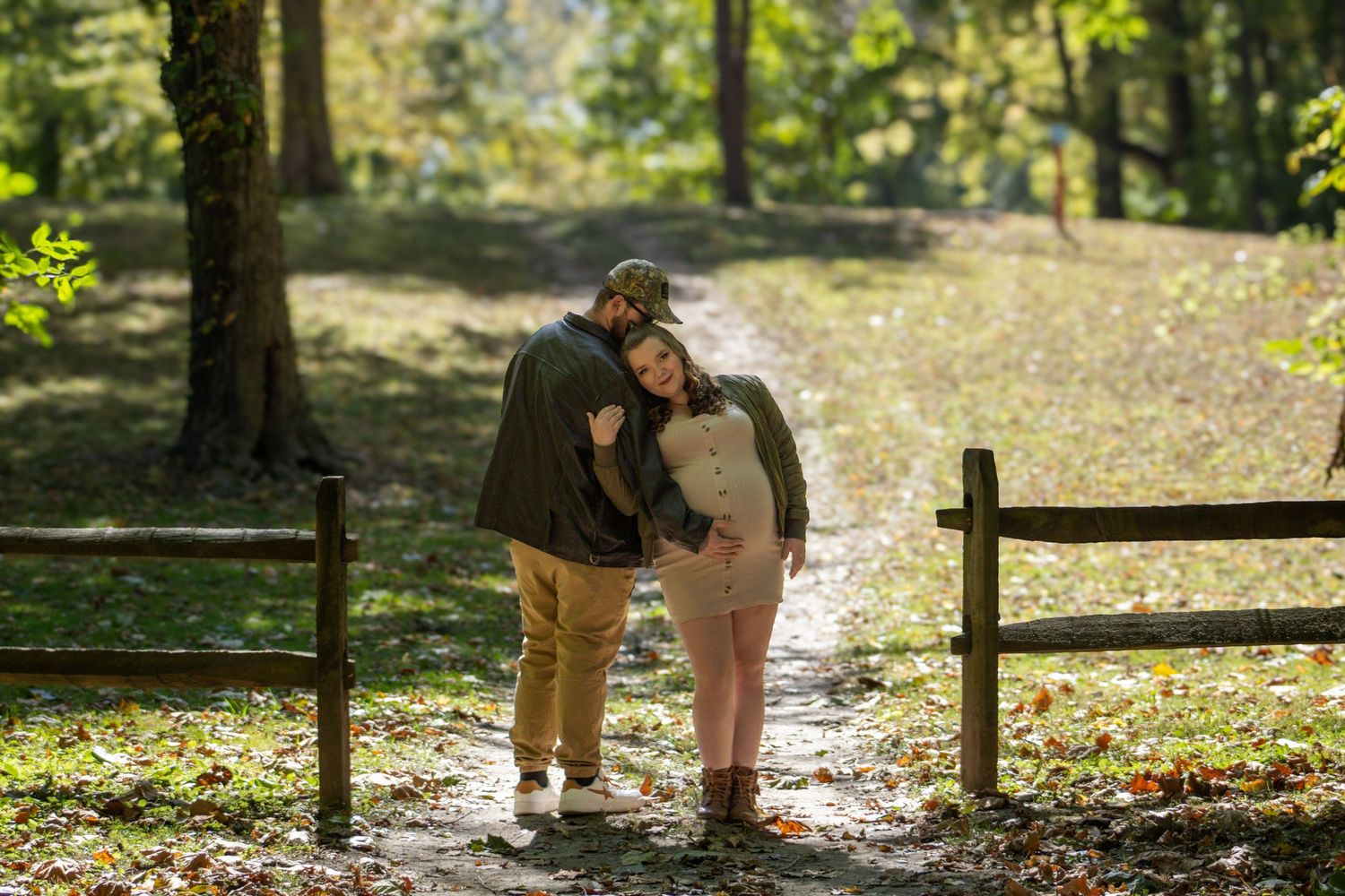 A couple embraces while standing on a sunlit forest path next to a wooden fence during autumn.