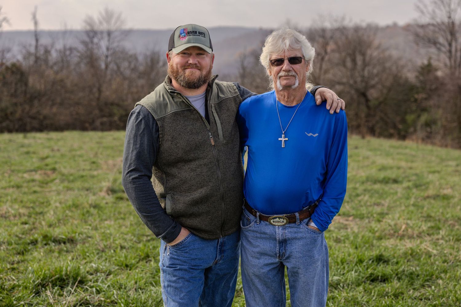 Two people stand together in a grassy field on a cloudy day, wearing casual attire including jeans and vests.