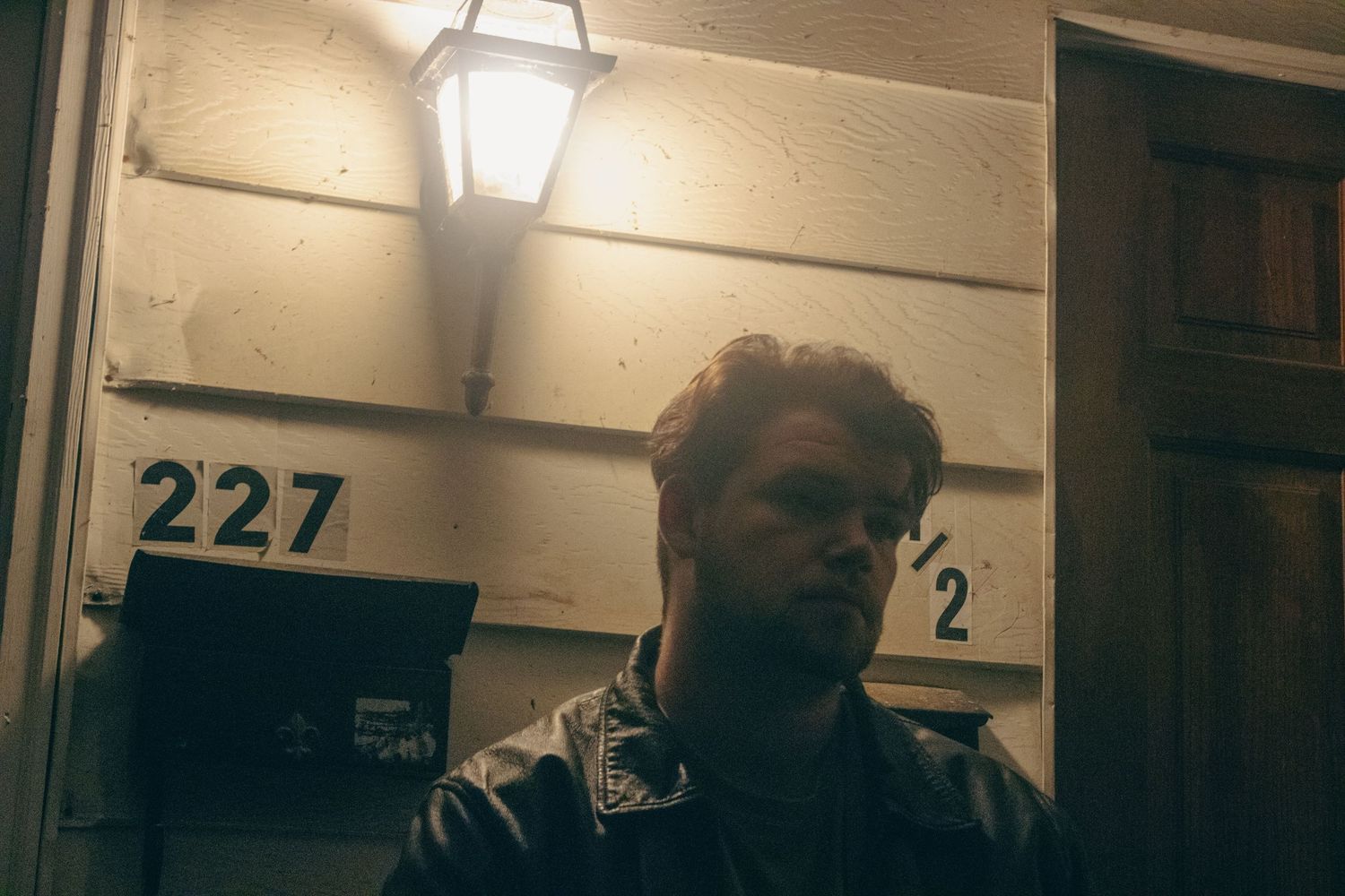 Two people sit in a dimly lit room with number 227 visible on the wall in the background.