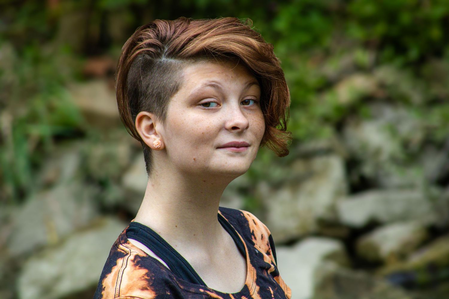 A person with an asymmetrical brown hairstyle wearing a floral top poses outdoors against a natural background.