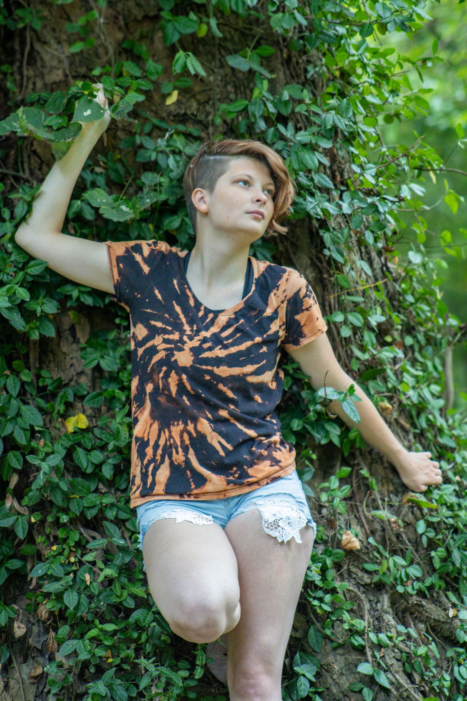 A person in a tie-dye shirt and white shorts poses playfully in front of green foliage.