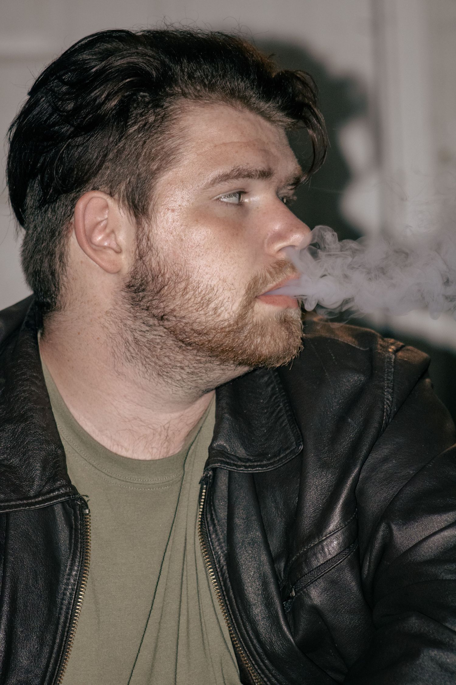 A person wearing a black leather jacket and olive shirt exhales smoke in a series of profile shots.