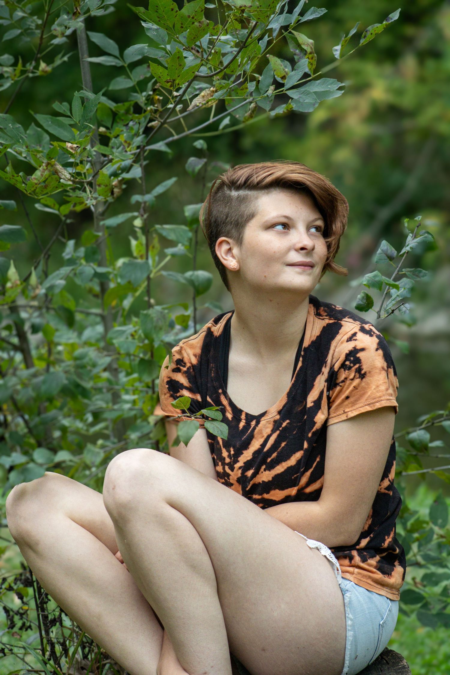 A person in an orange tie-dye shirt sits among green foliage in a garden setting.