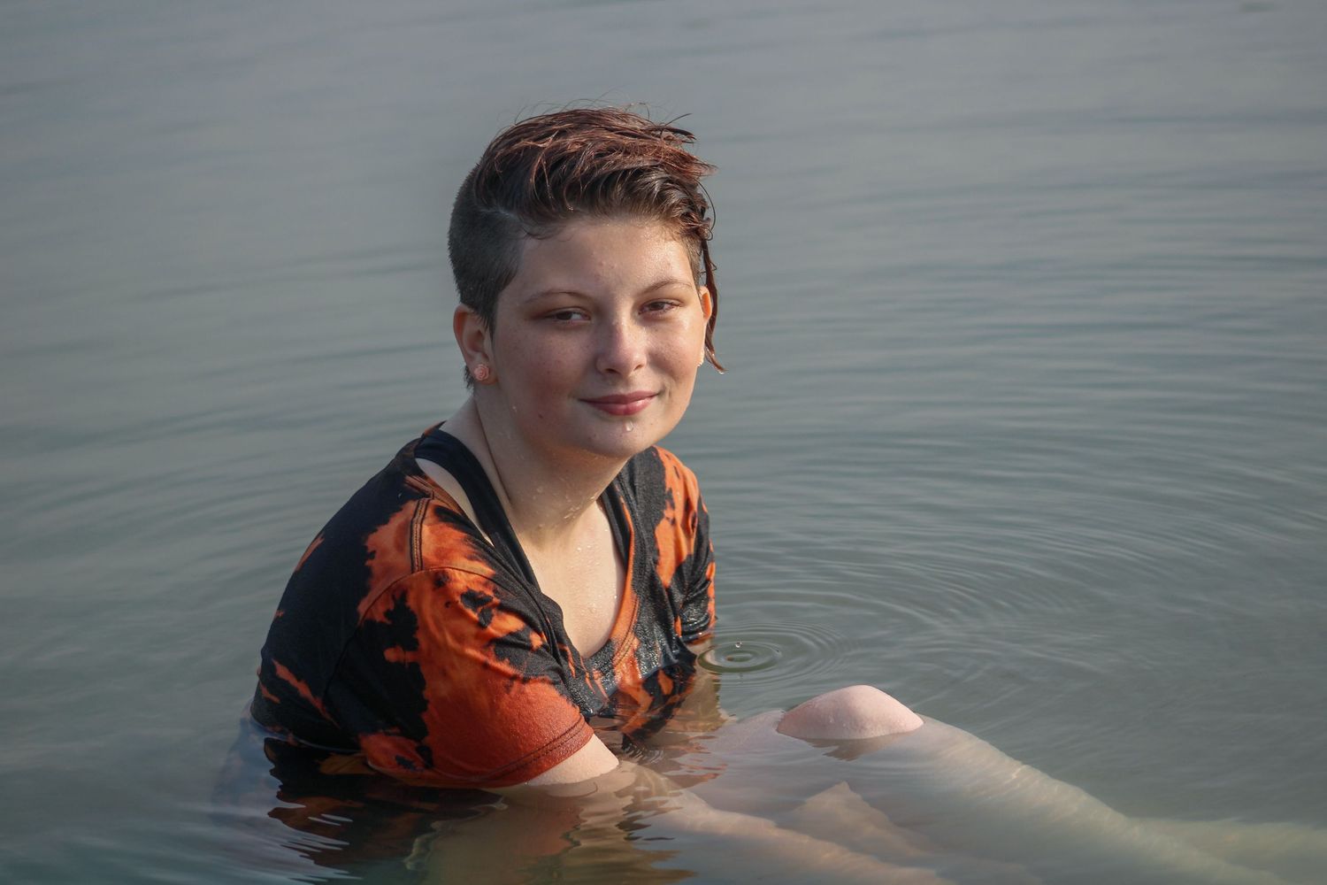 A person wearing an orange life vest swims in calm, gray water on an overcast day.