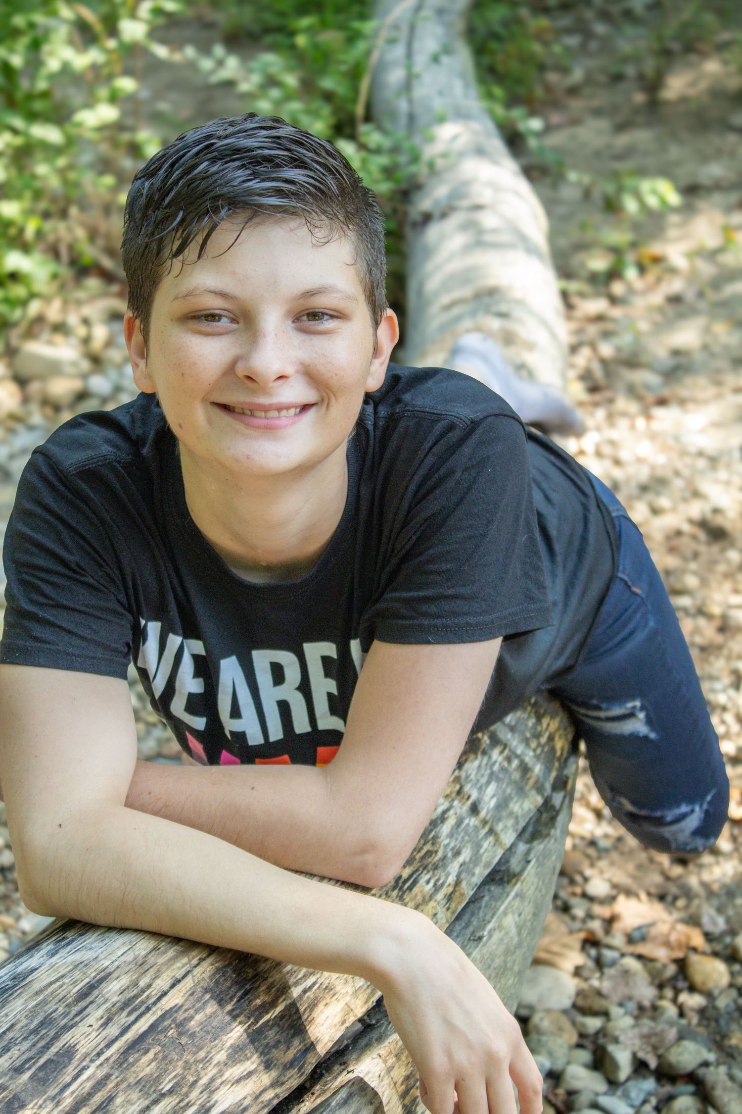 Someone in a black t-shirt with short dark hair rests on a fallen log in a wooded outdoor setting.