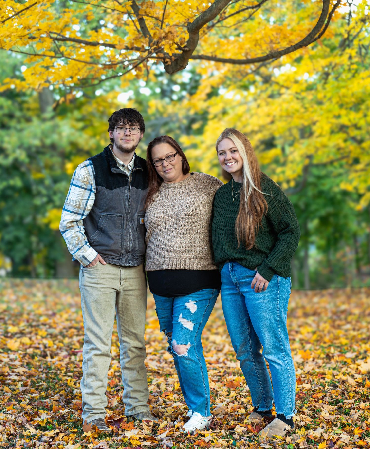 Three people stand close together on fallen autumn leaves with vibrant yellow foliage in the background.