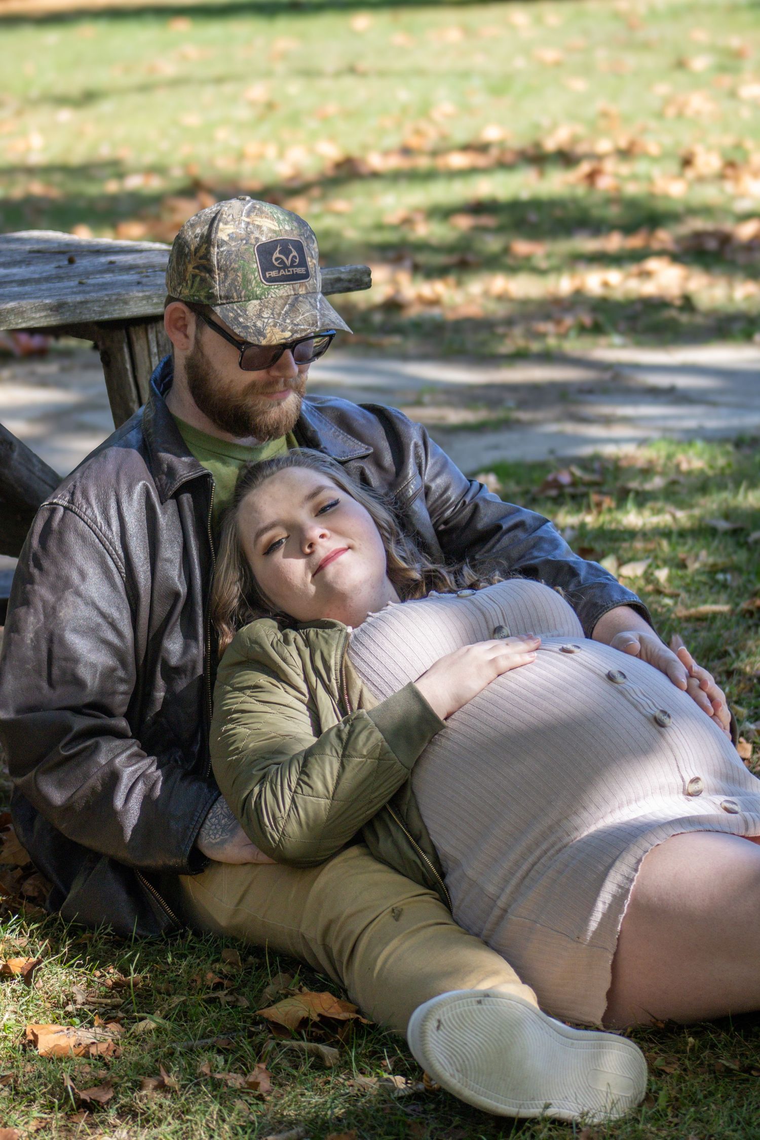 A couple rests together on grass during a peaceful autumn day with fallen leaves scattered around them.