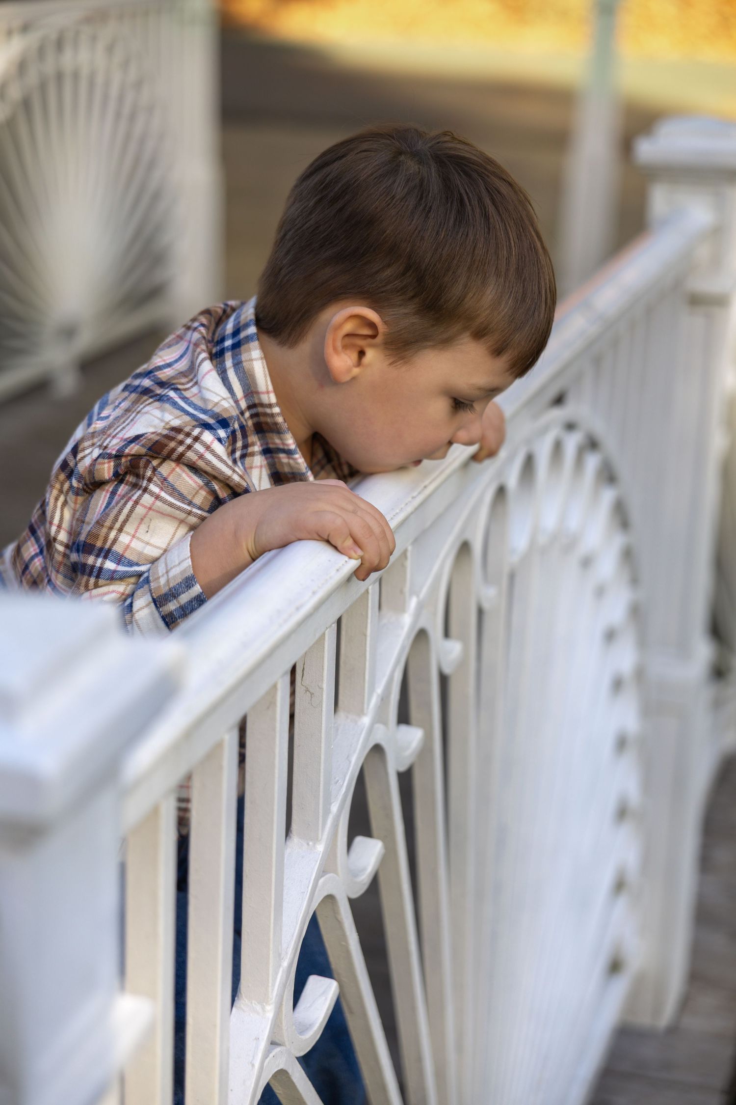 A child in a plaid shirt leans over a white porch railing during a golden sunset.