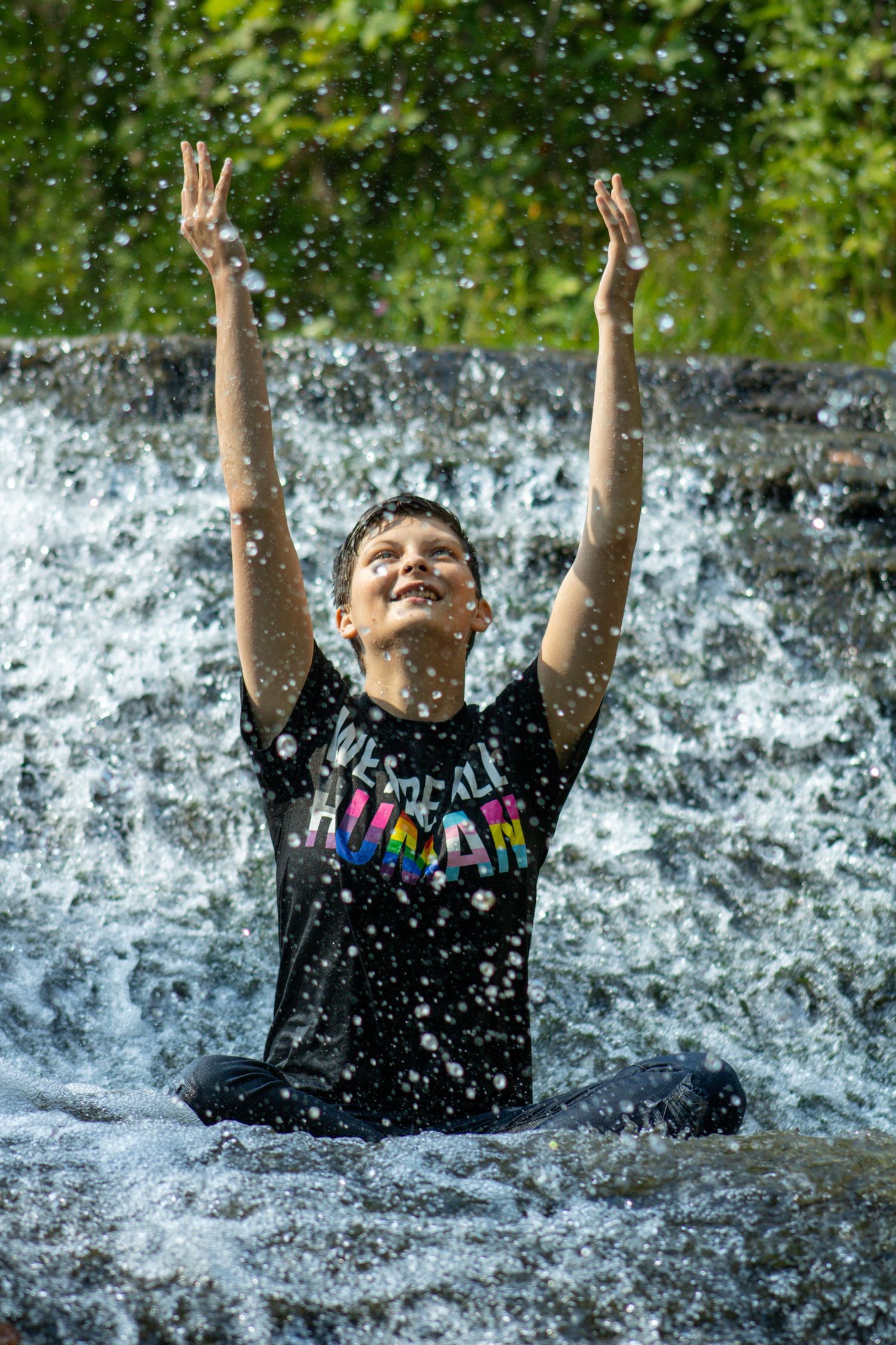 Someone in a colorful t-shirt raises their arms joyfully while playing in a cascading waterfall on a sunny day.