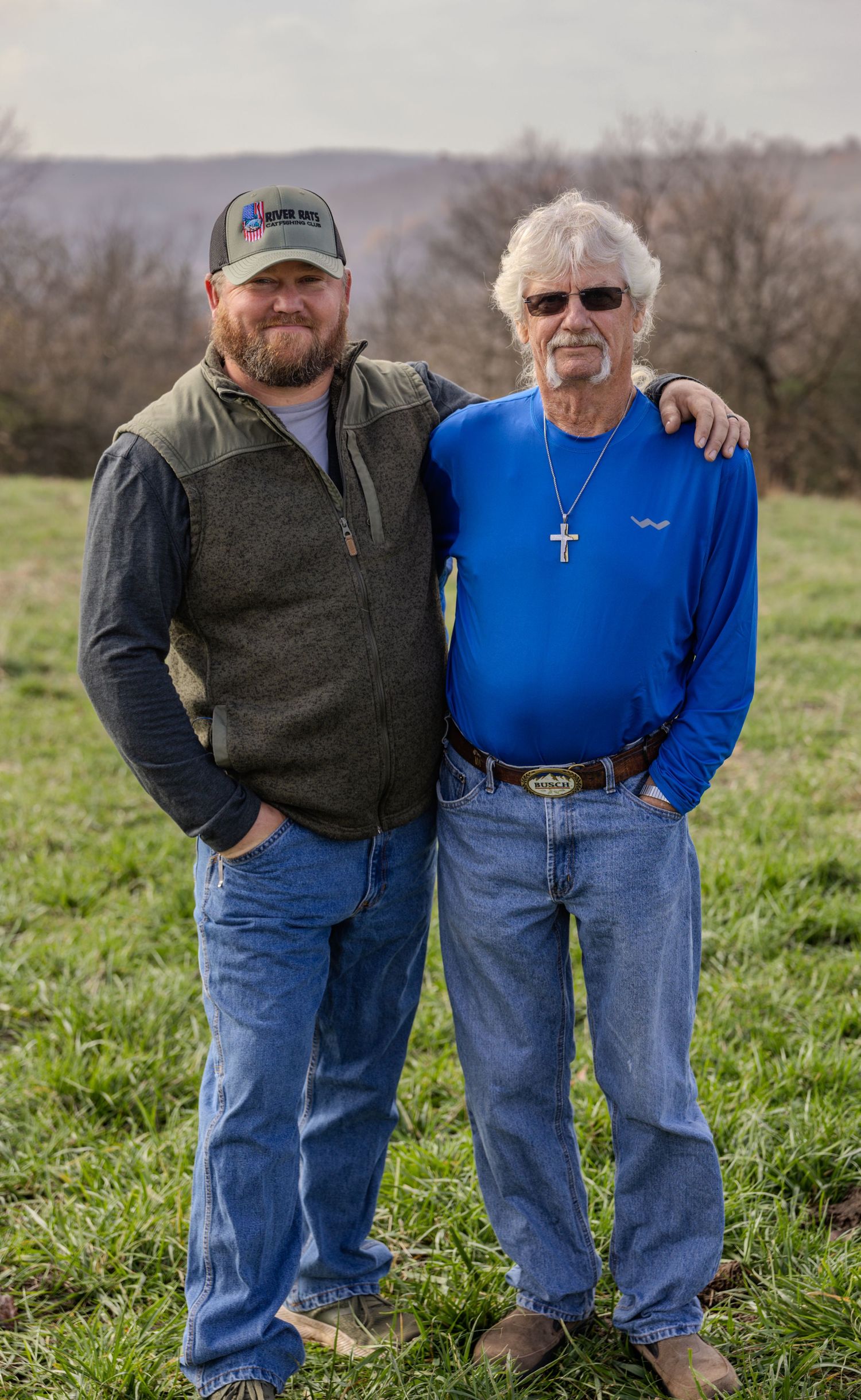 Two people wearing jeans and sweaters stand together in a grassy field on an overcast day.