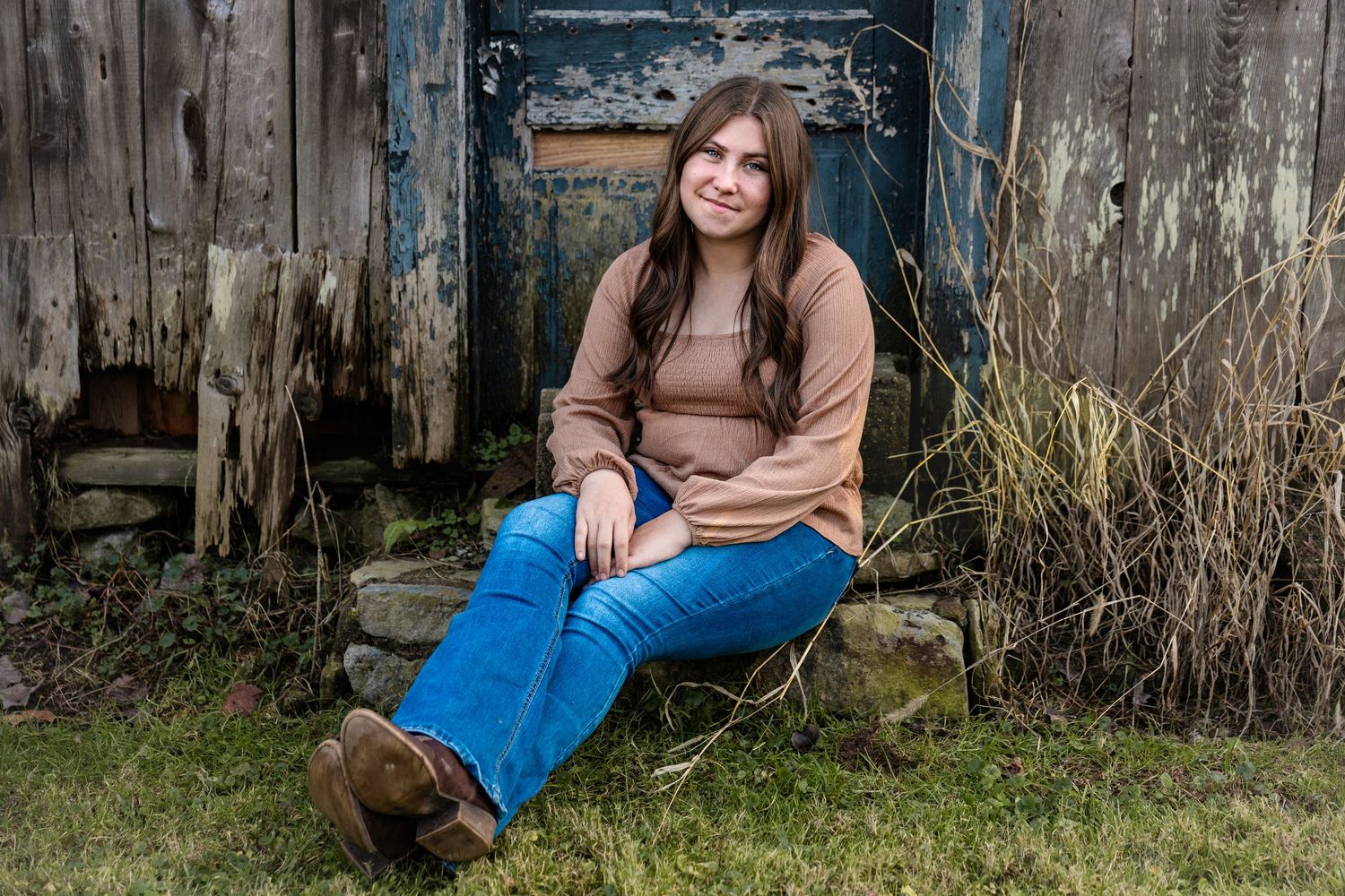 A person sits outdoors in brown clothing against a rustic wooden fence with natural foliage in the background.