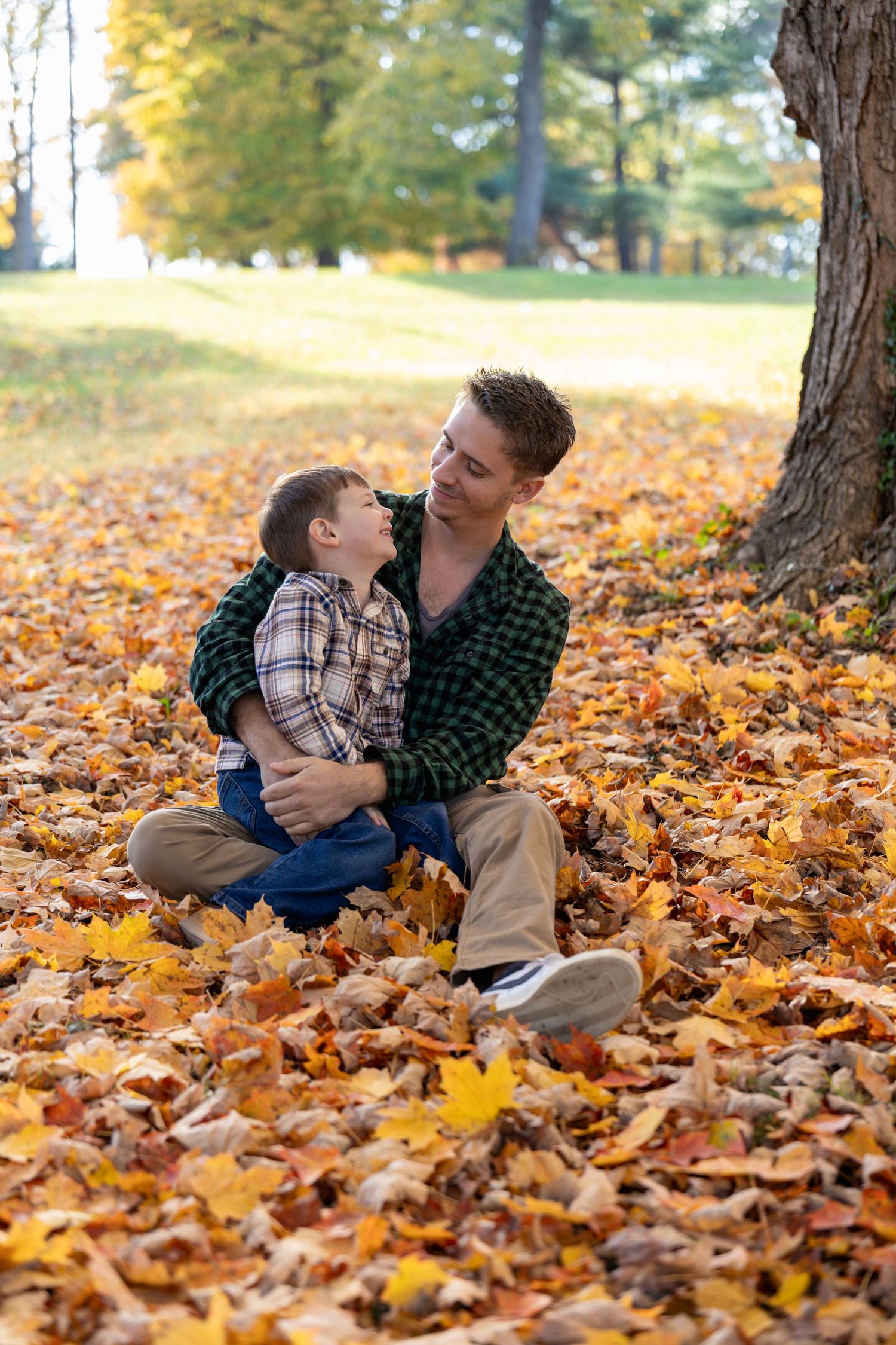 Two people in plaid shirts share a tender moment while sitting on fallen autumn leaves in a park.