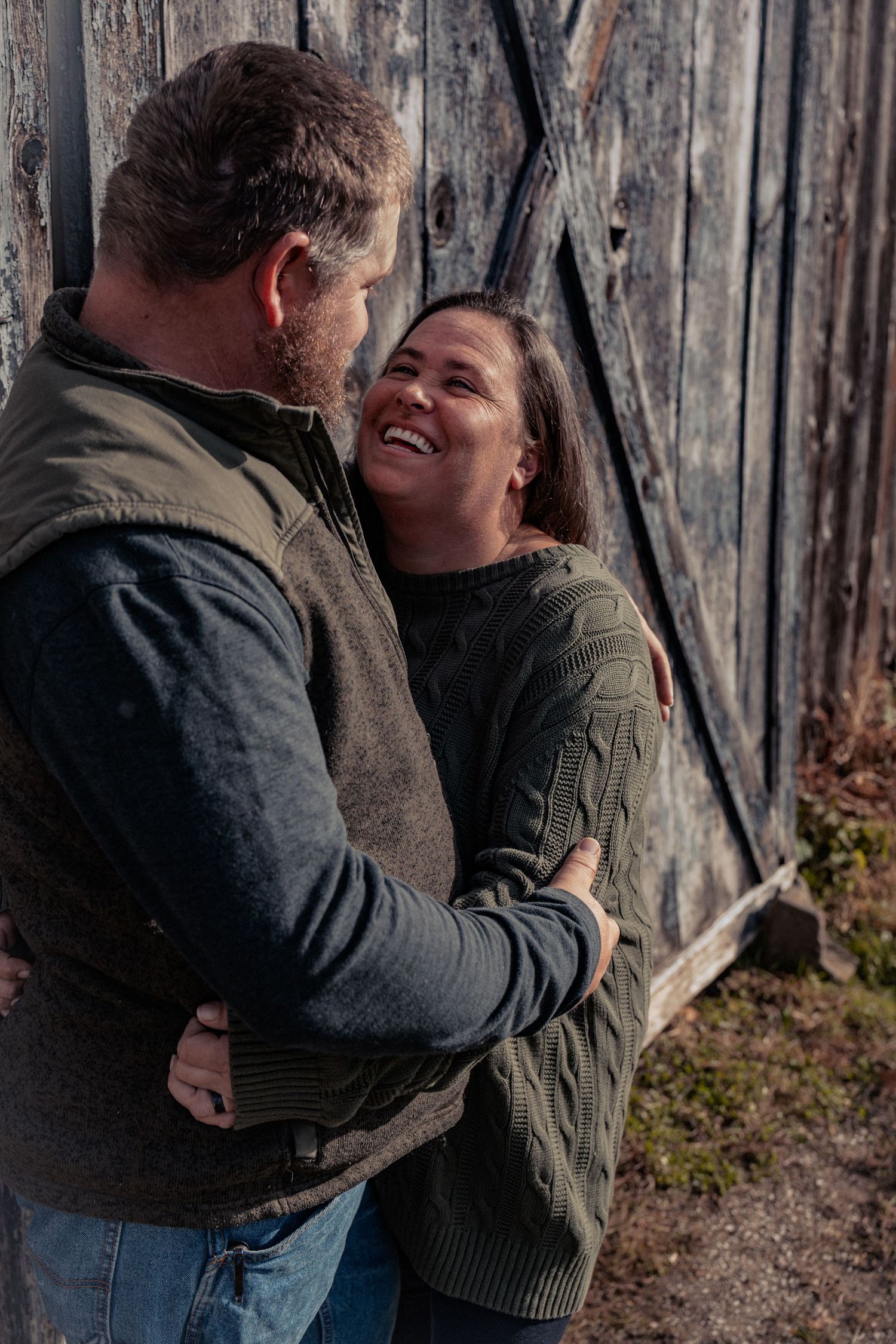 A couple shares a tender embrace and laugh together against a rustic wooden wall background.