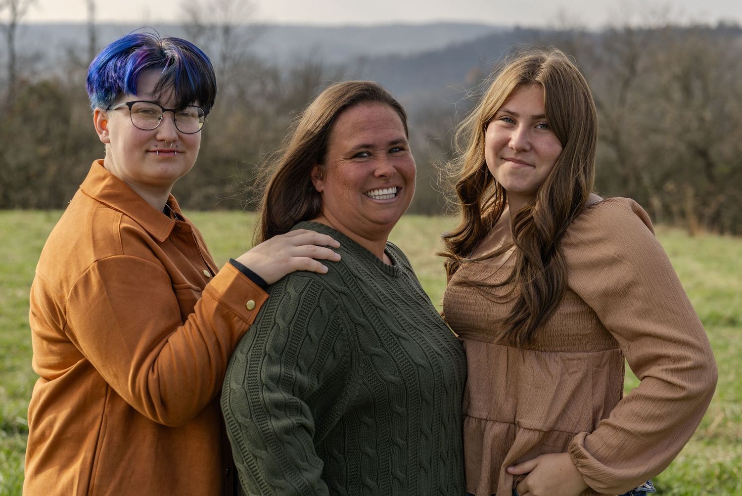 A group of people wearing warm coats embrace and laugh together outdoors in a natural setting during autumn.