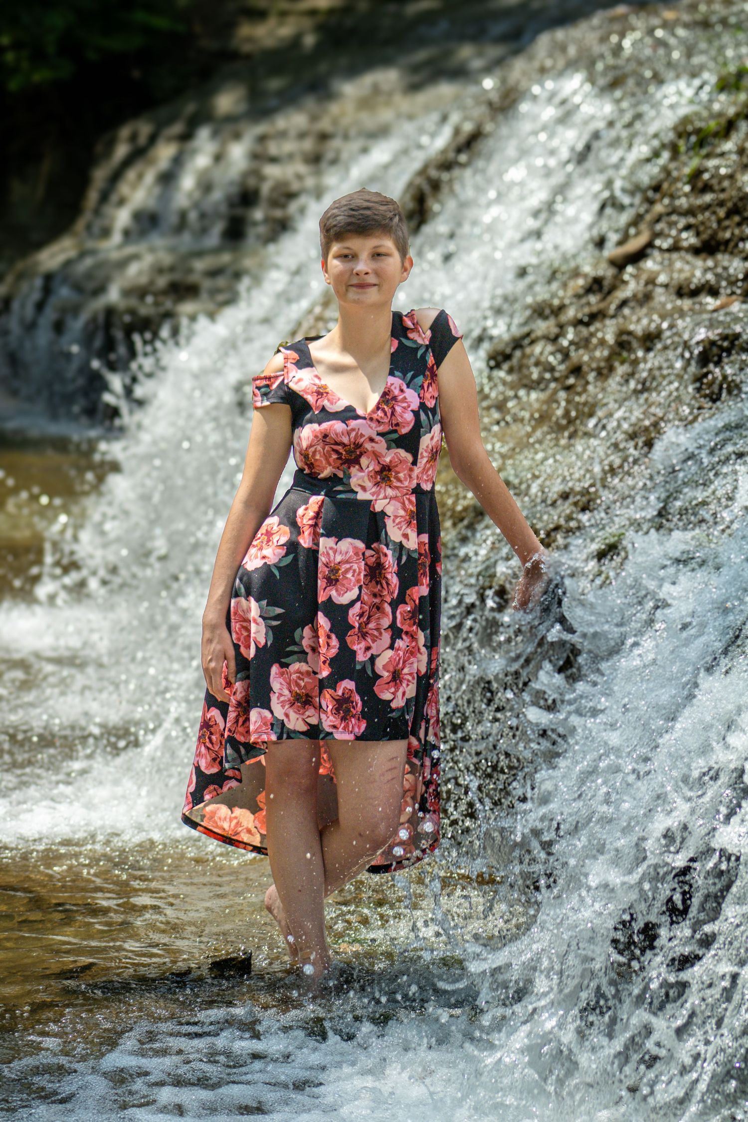 A floral dress-clad figure stands in flowing water at the base of a cascading waterfall on a sunny day.