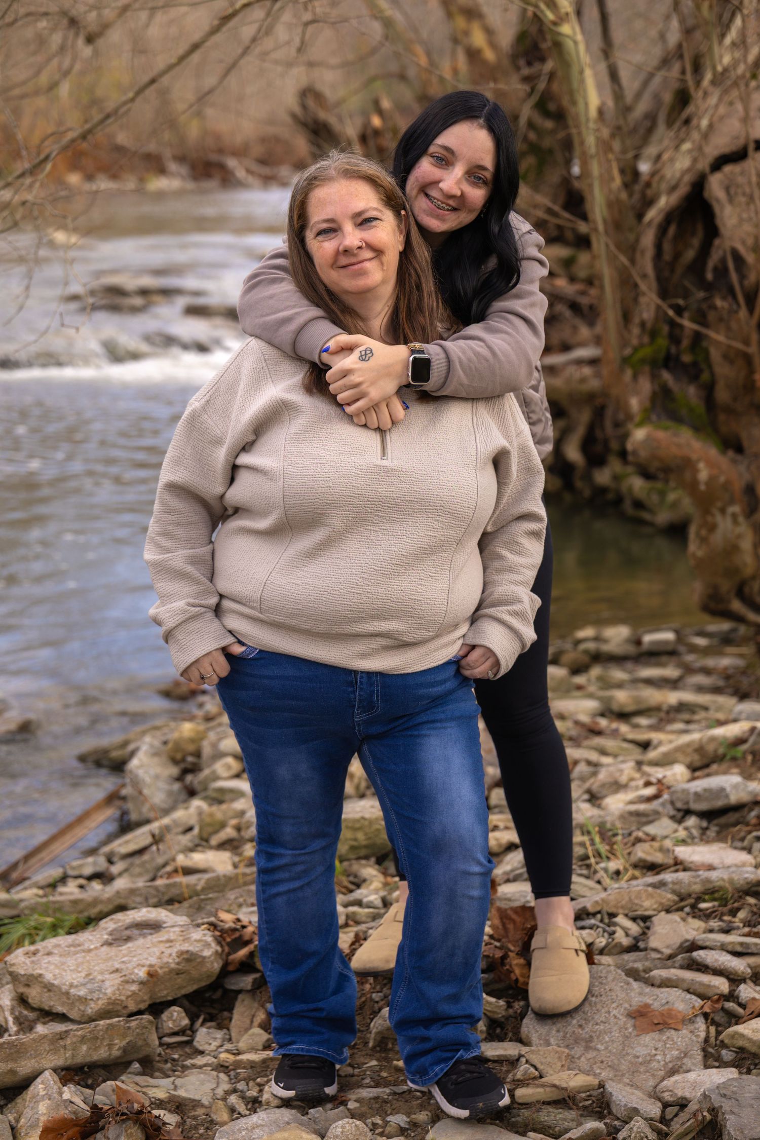 Two people embrace while sitting on rocky terrain near a flowing stream in an outdoor nature setting.