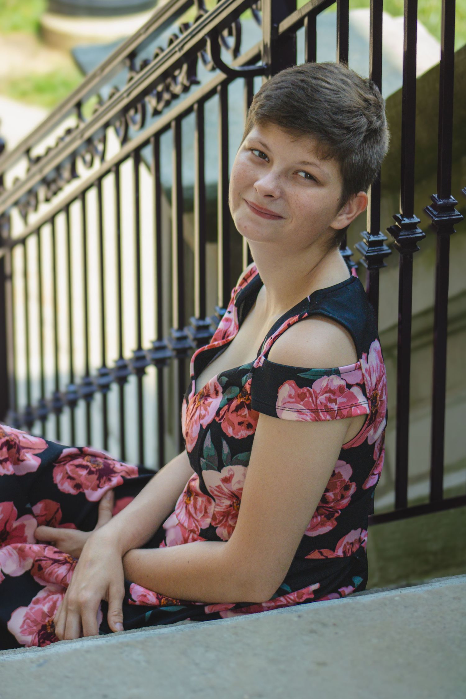 A person wearing a black and pink floral dress sits on outdoor steps near a decorative metal railing.