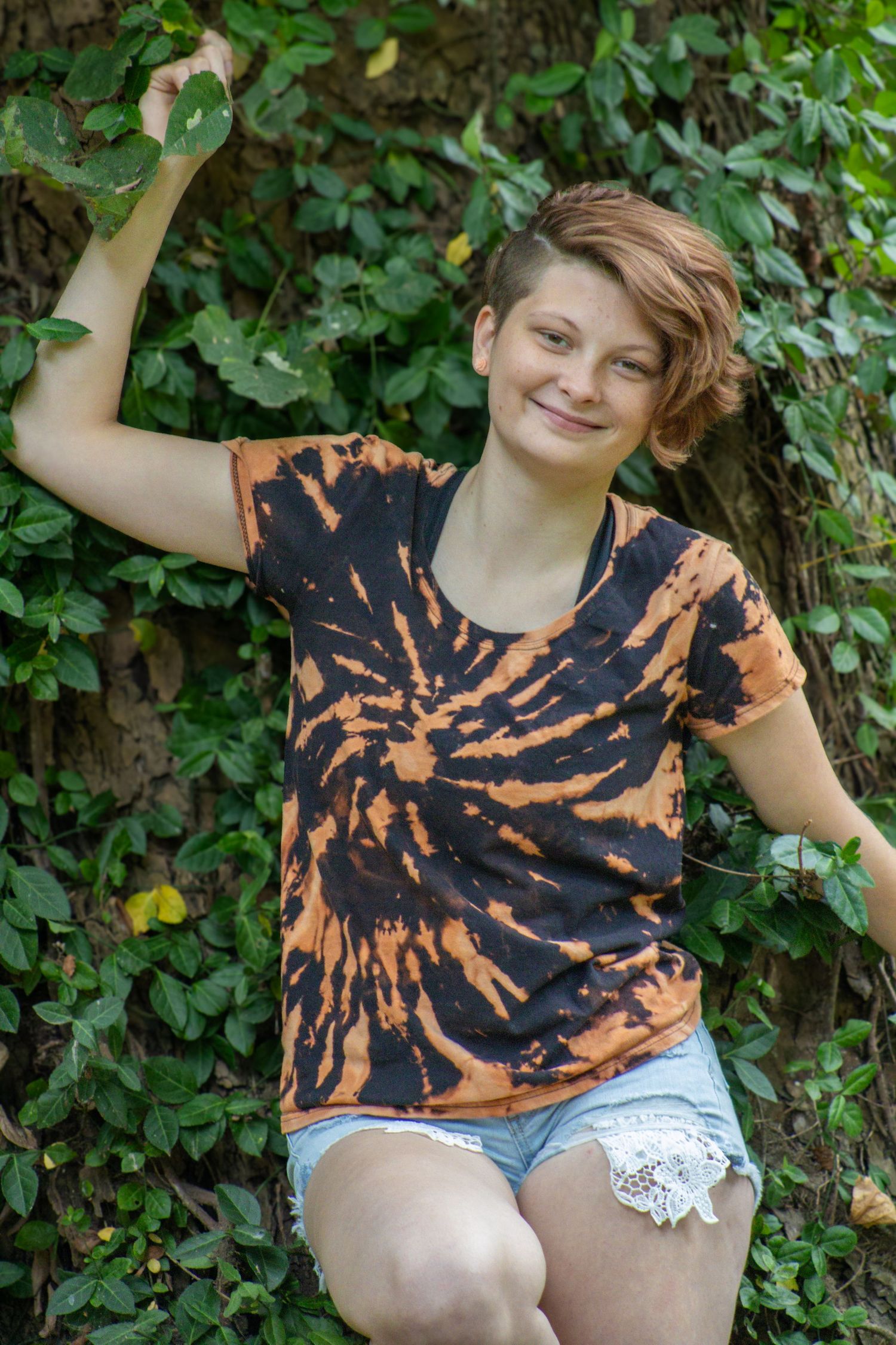 A person wearing a black and orange tie-dye shirt and light colored shorts poses in front of green foliage.