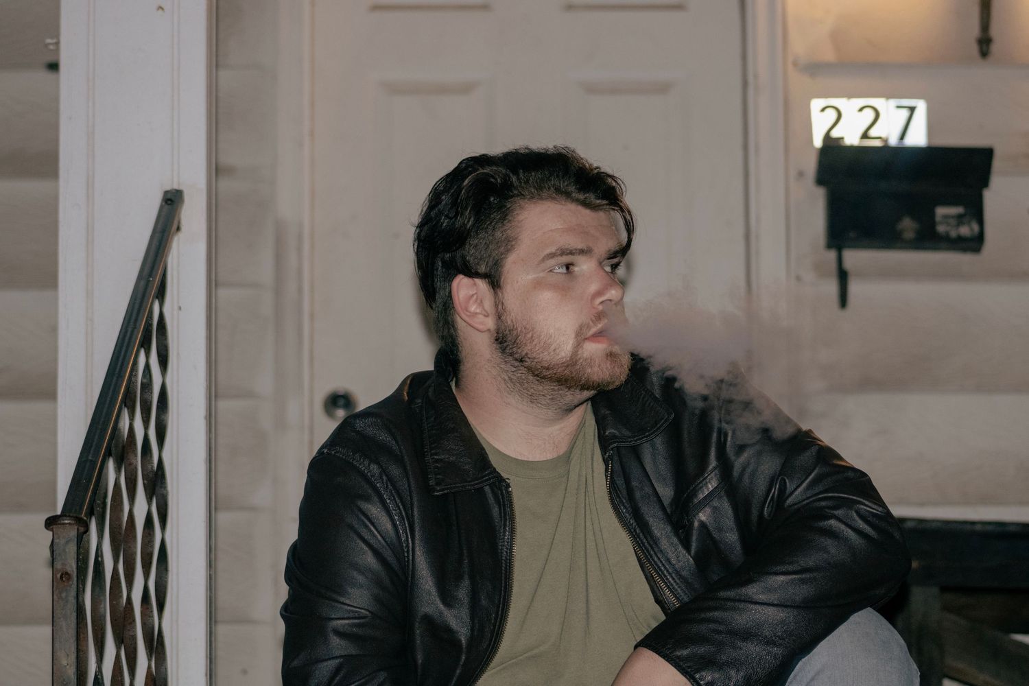 Person in dark jacket sits on porch steps in evening lighting with house number visible.