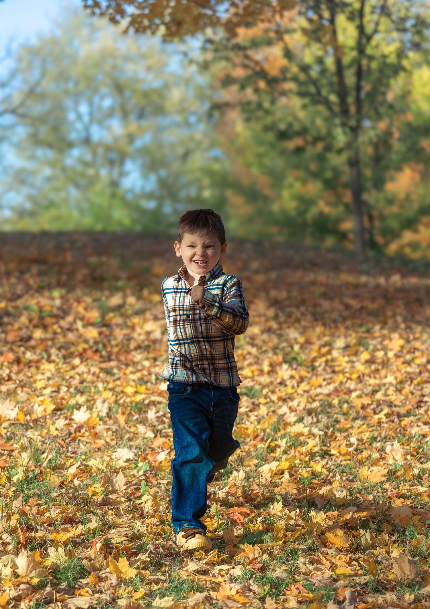 A child in a plaid shirt and jeans runs joyfully through fallen autumn leaves on a sunny day in the park.