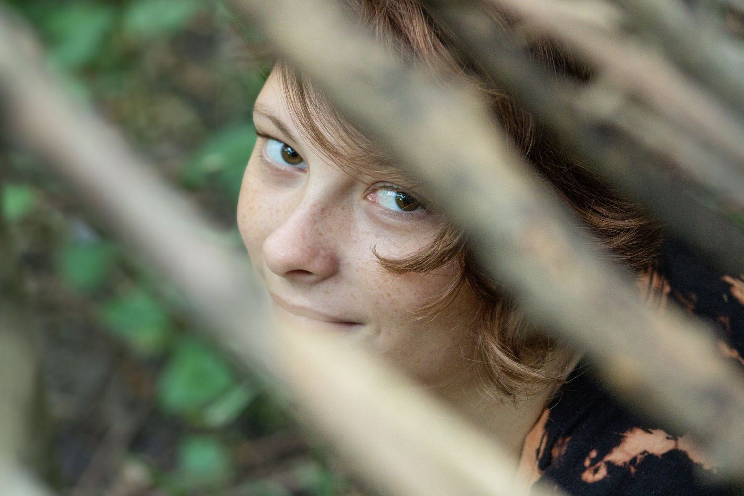 A person peeks playfully through tree branches and leaves in an outdoor setting.