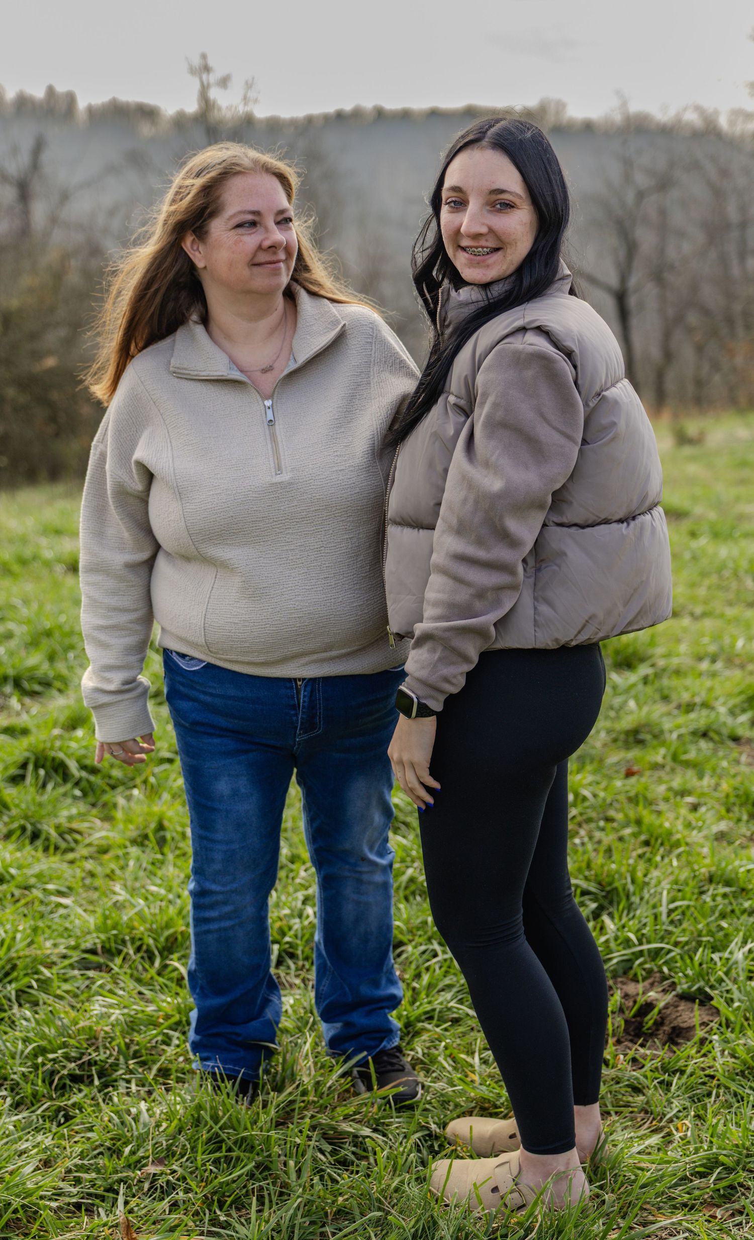 Two people in casual clothing stand together on a grassy hillside on an overcast day.
