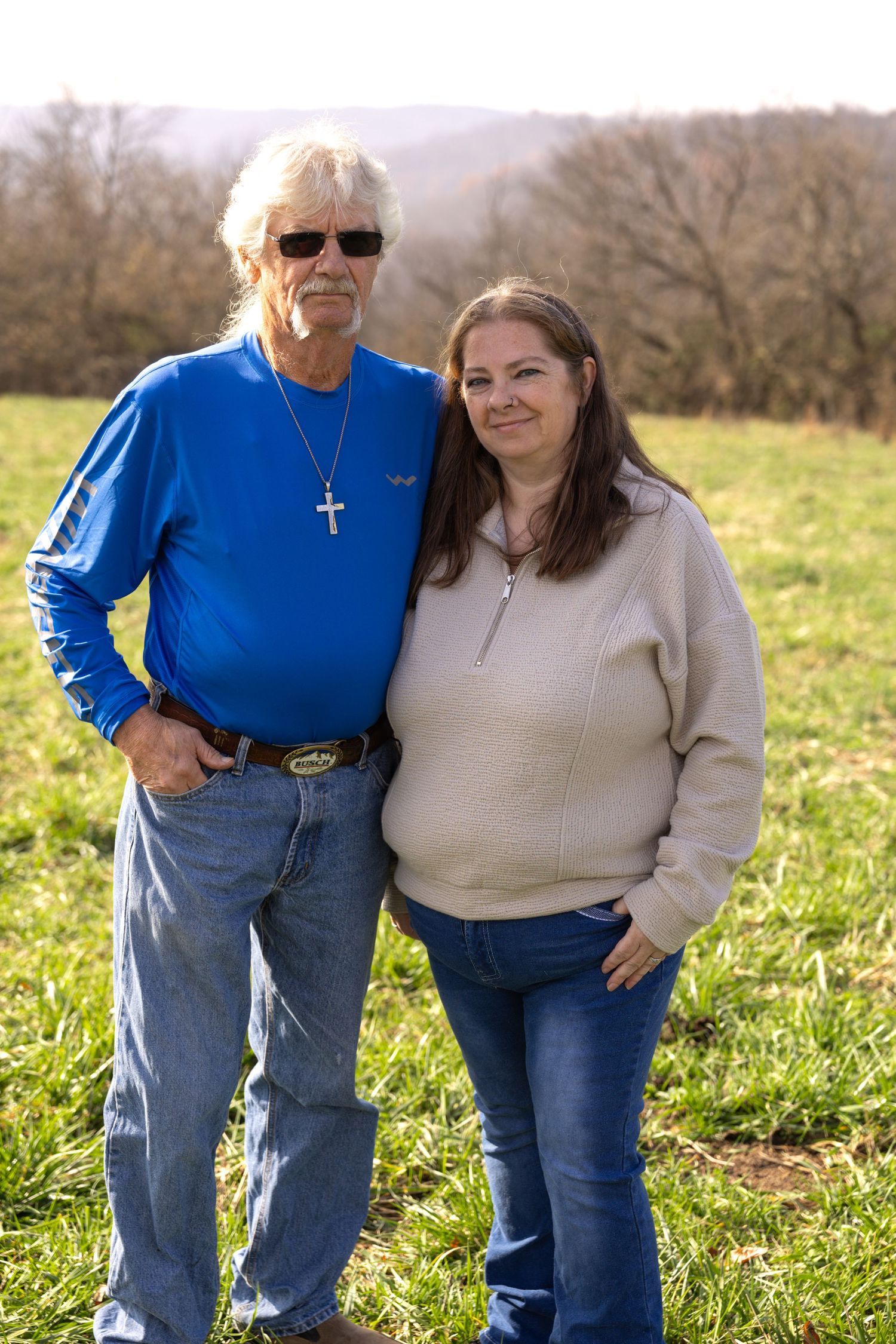 Two people stand together in a grassy field on a sunny autumn day wearing blue and beige casual clothing.