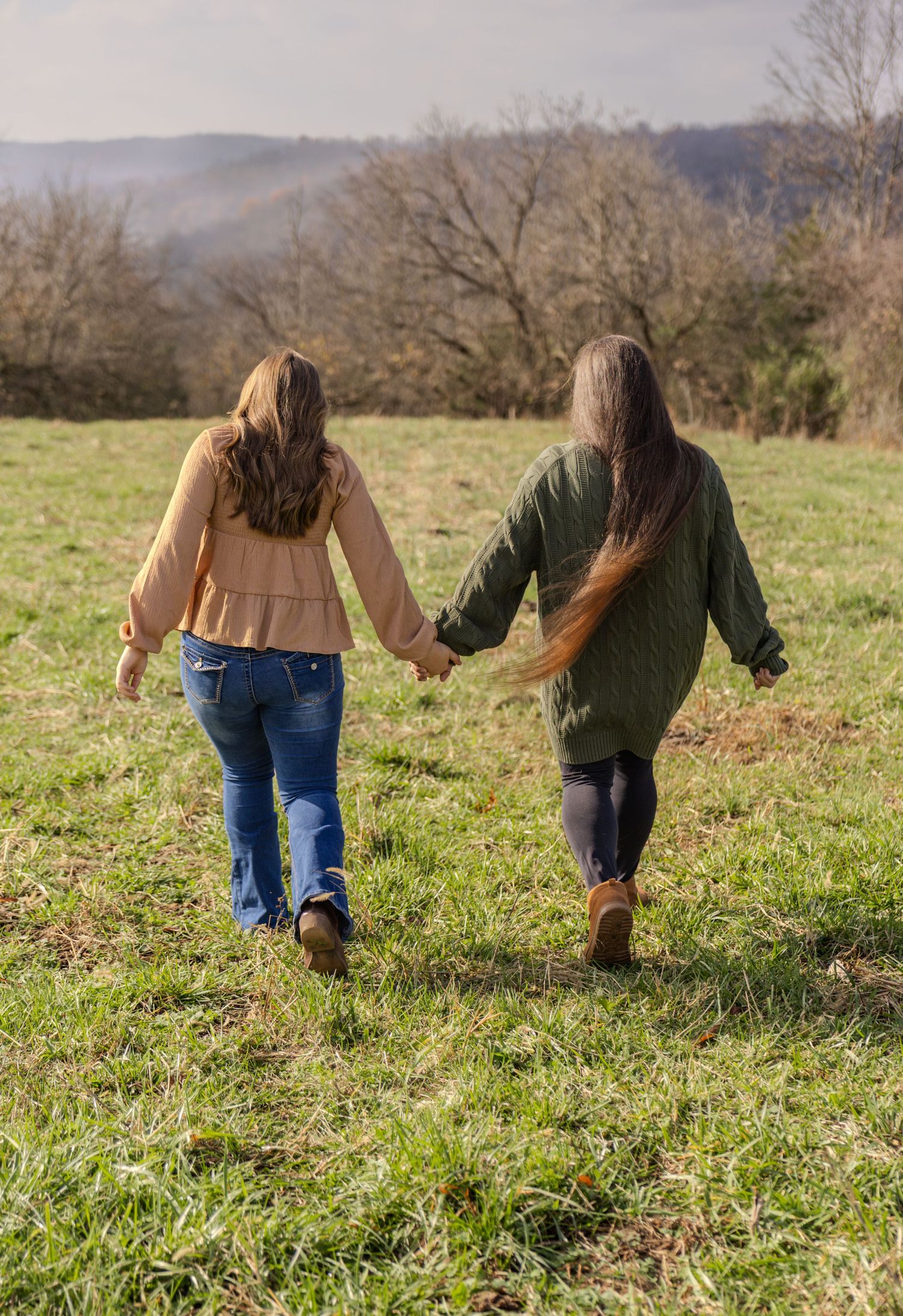 Two people holding hands while walking through a grassy field on a sunny day with hills in the background.