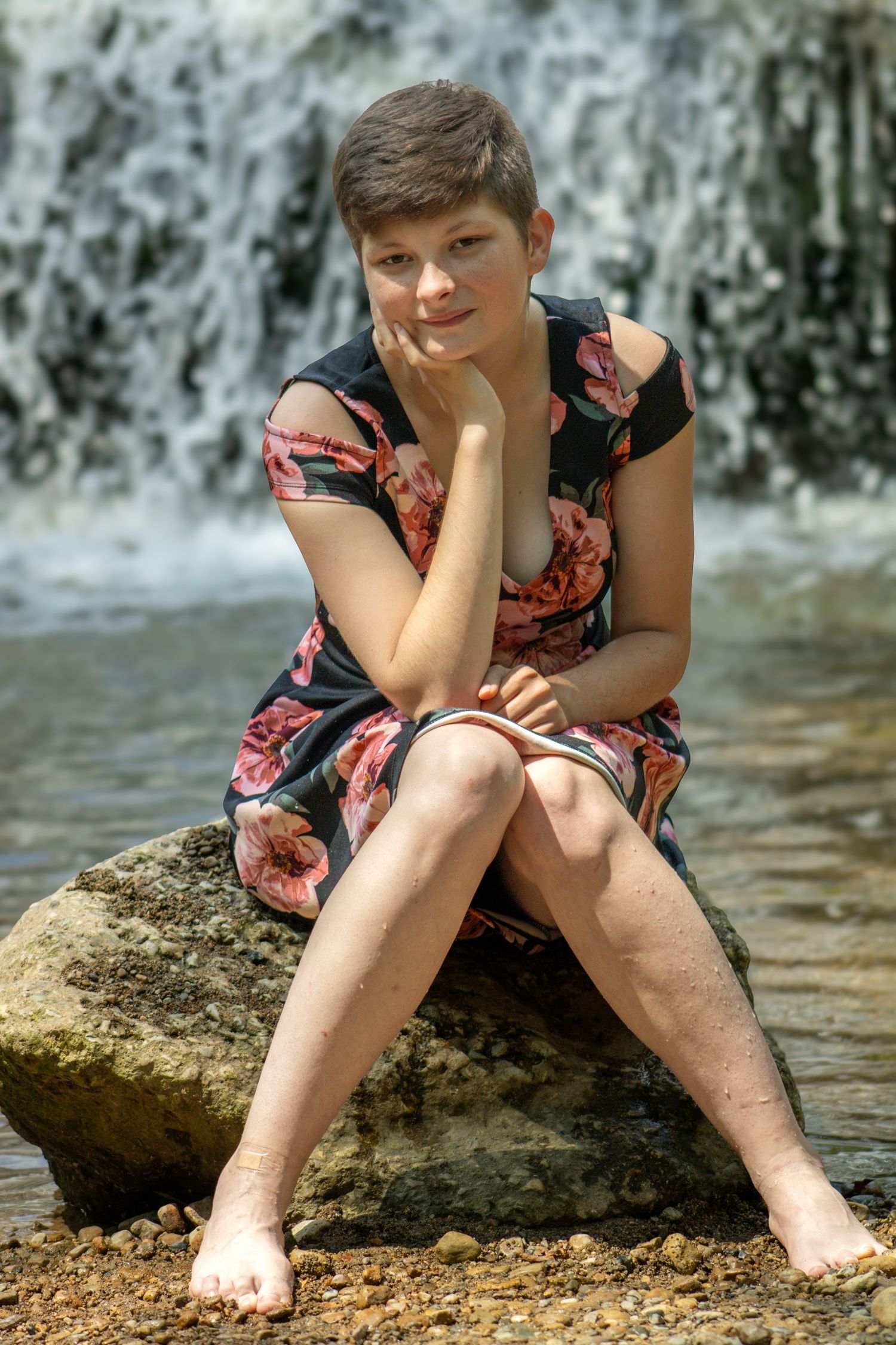 Someone sits on rocks by a waterfall wearing a floral dress on a sunny day.