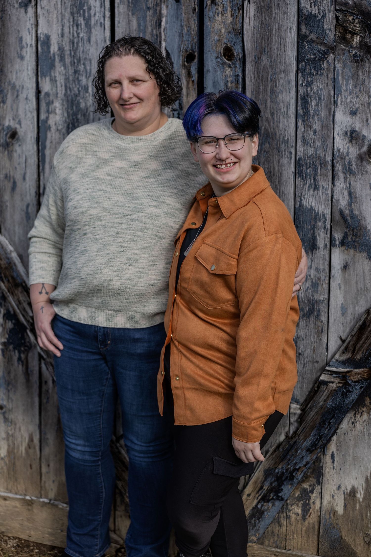 Two people pose together against a weathered wooden wall in casual attire including jeans and a brown jacket.