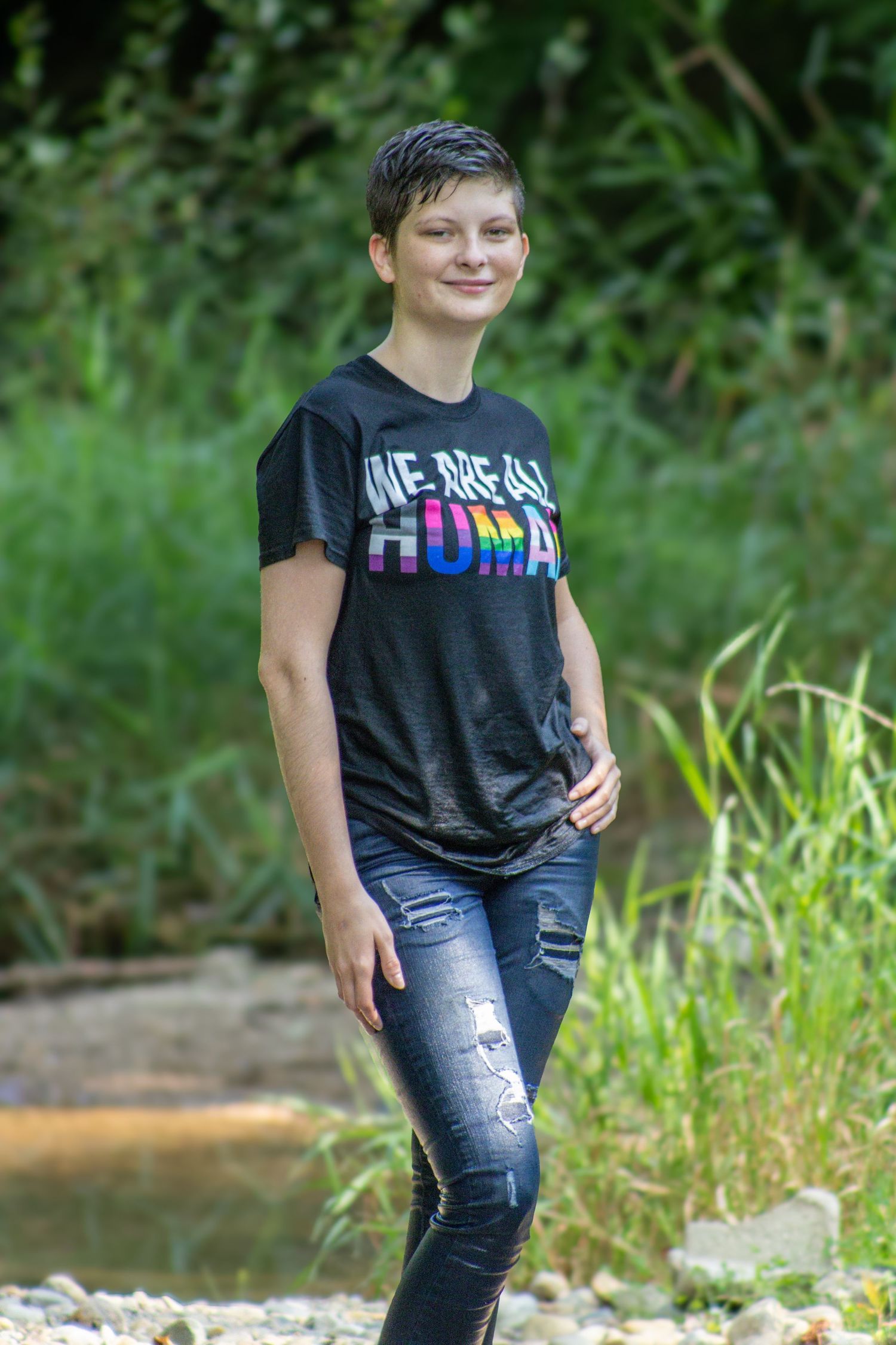 A person in ripped jeans and a dark t-shirt poses casually near a stream in a natural outdoor setting.