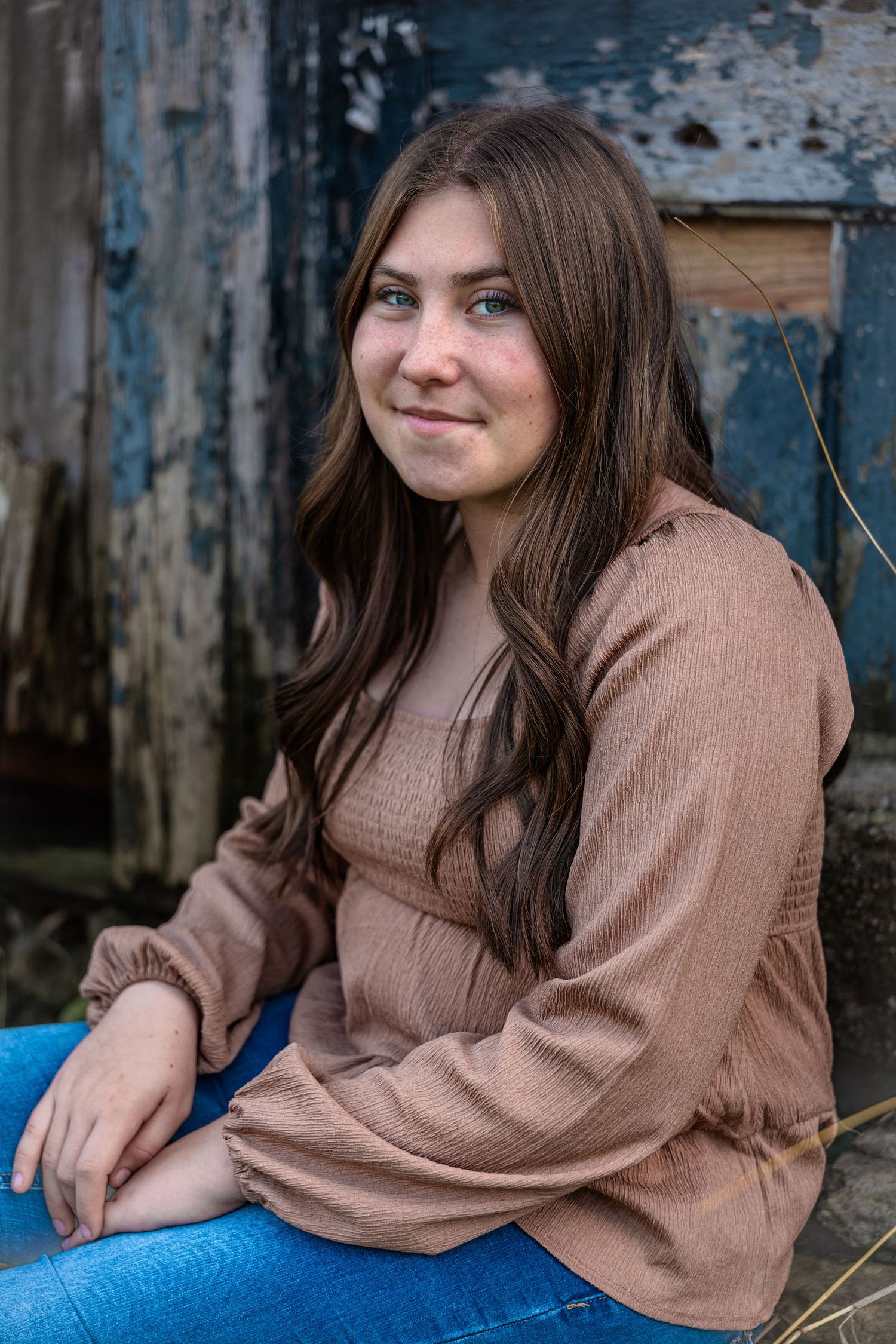 A person in a brown sweater and jeans sits casually outdoors against a weathered blue background.