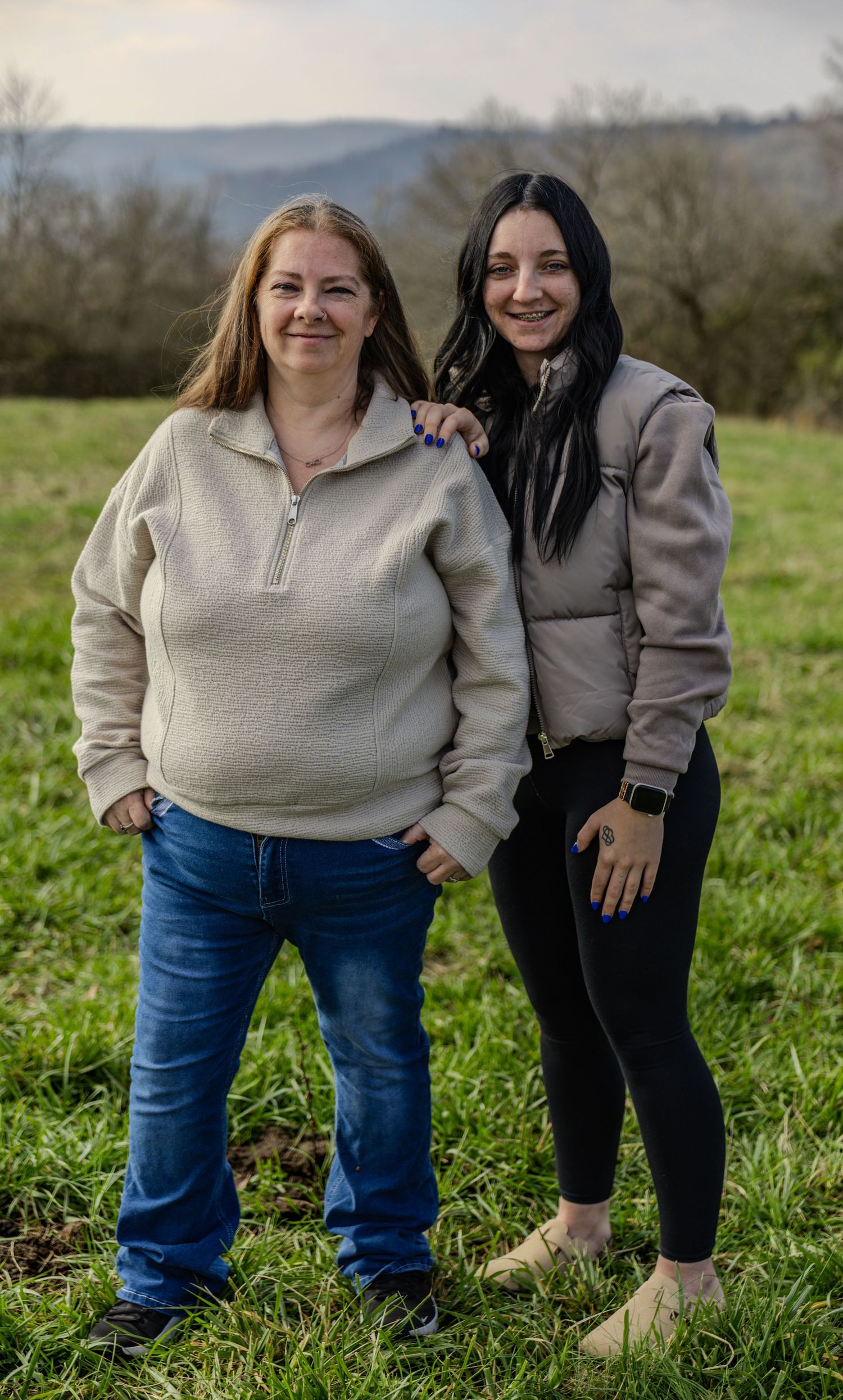 Two people in casual sweaters and pants stand together in a grassy field with rolling hills in the background.
