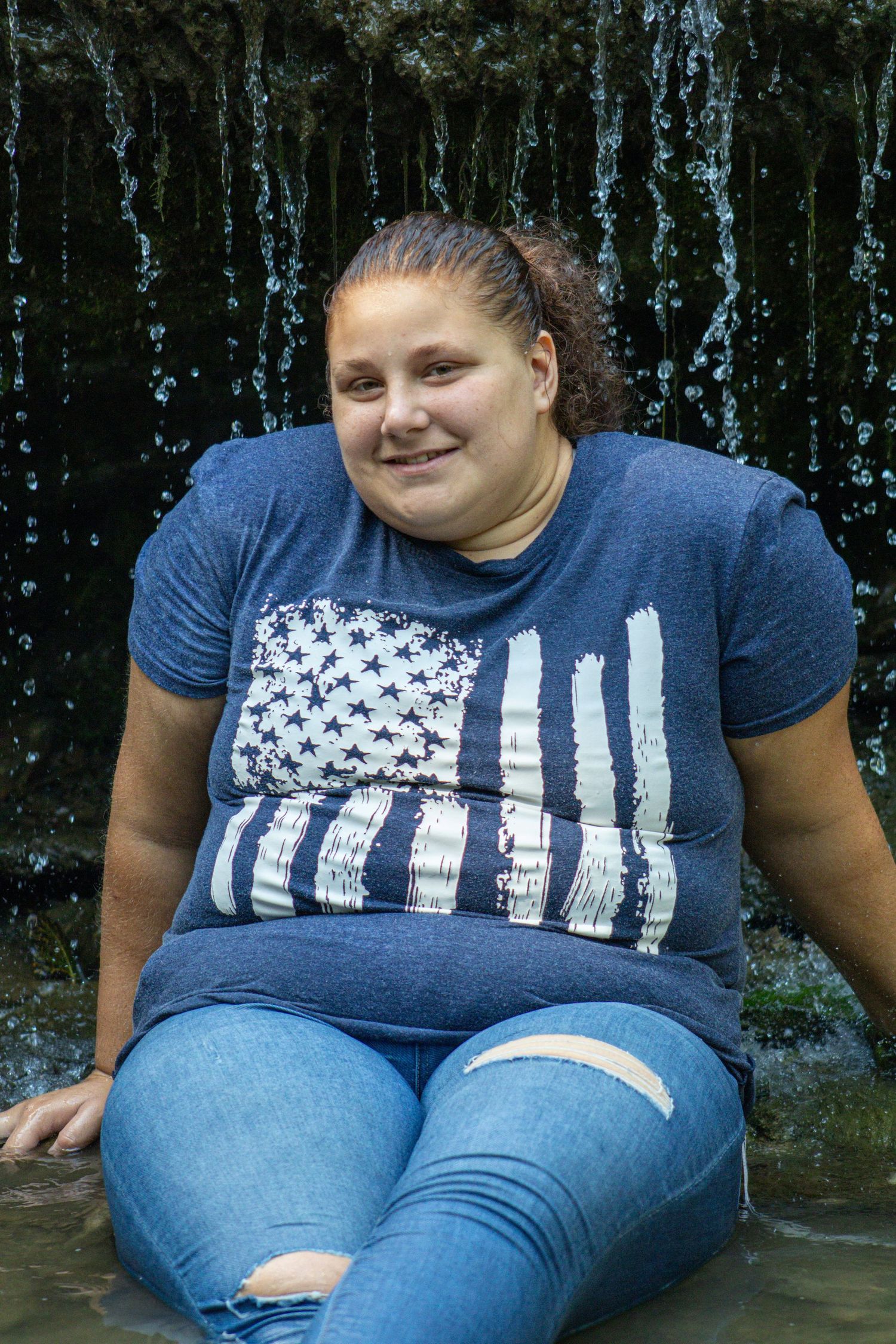 Person in a distressed American flag t-shirt and jeans sitting on a stone surface with a dark background.