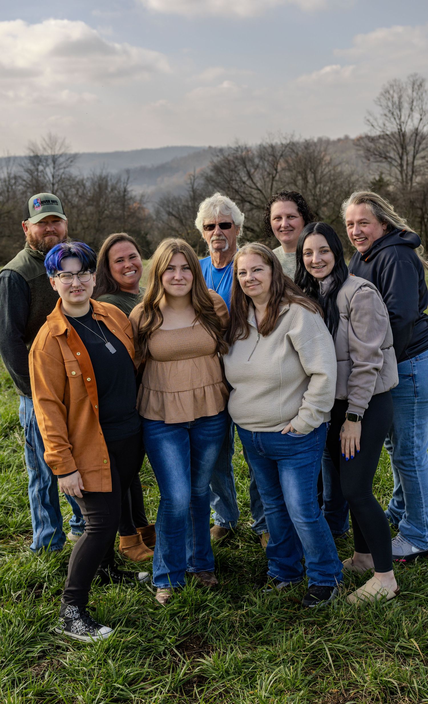 A group stands together on a grassy hillside with mountains and bare trees visible in the background on a cloudy day.