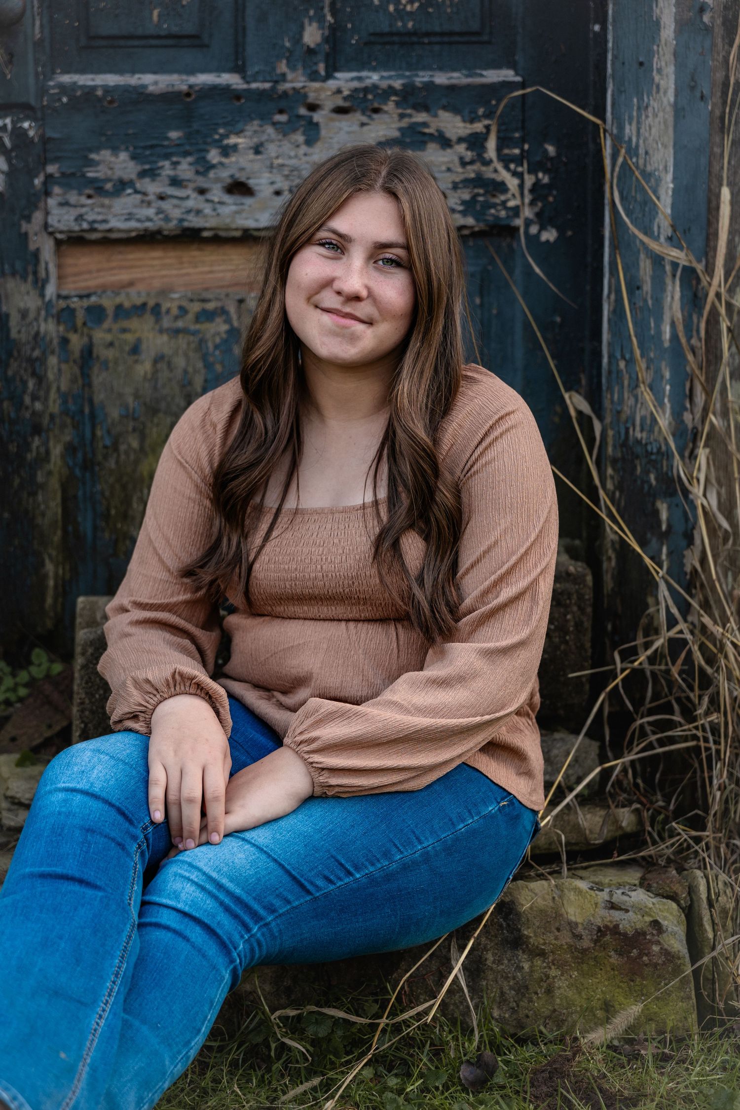 A person in a brown sweater and blue jeans sits casually against a weathered blue wall outdoors.