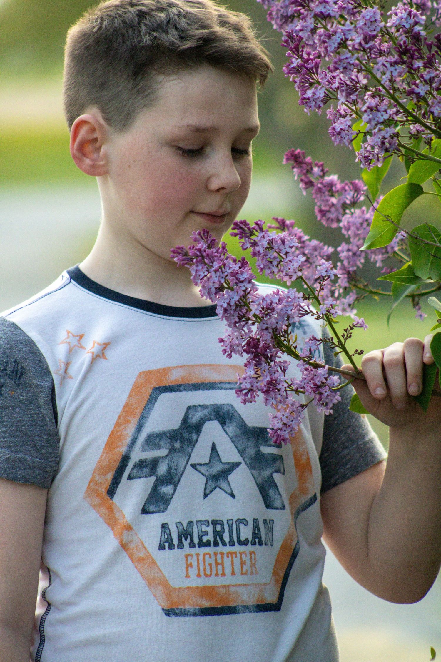 A child in a white American Fighter t-shirt smells blooming purple lilac flowers in a springtime garden setting.