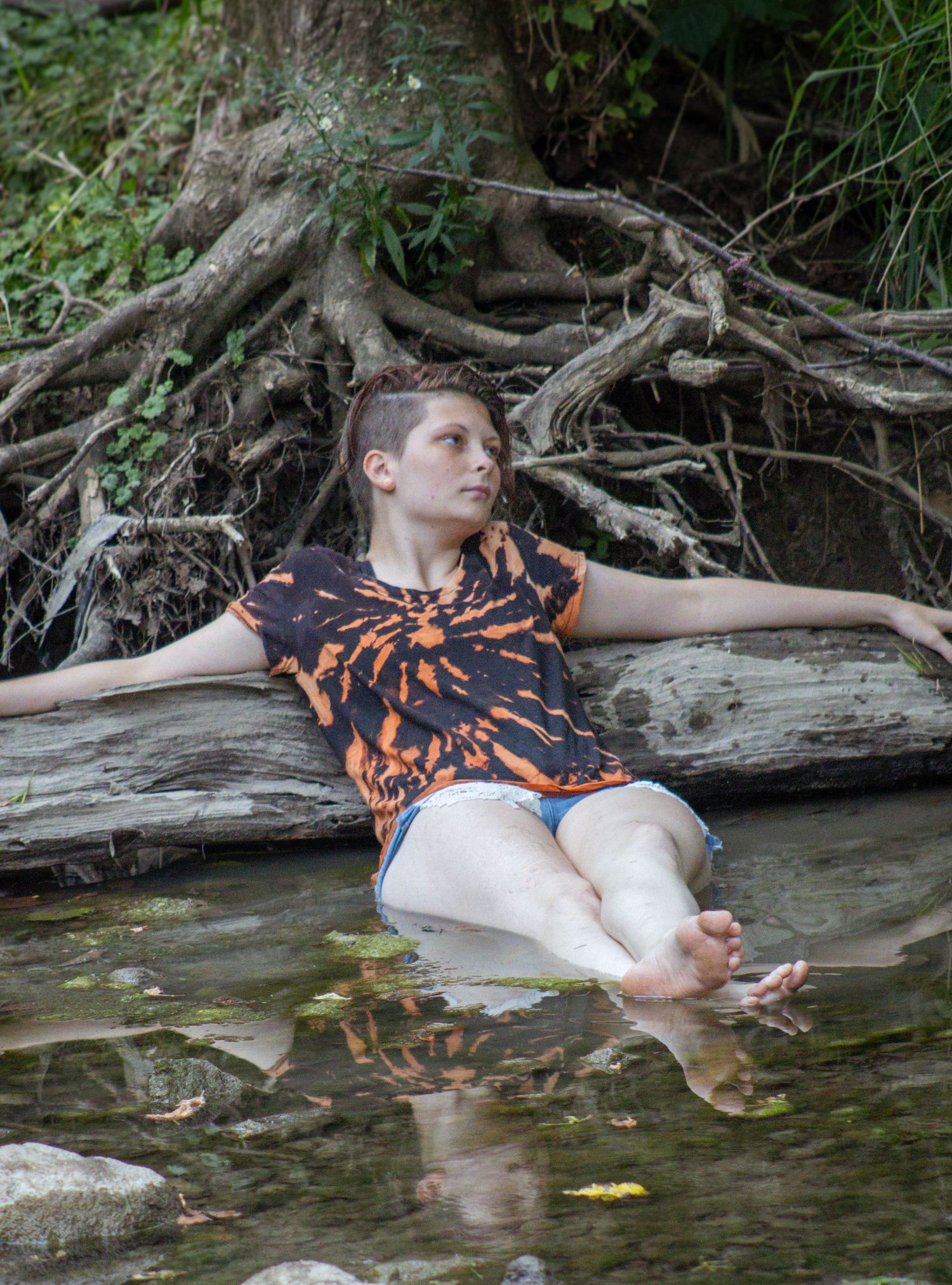 Someone in an orange and black patterned shirt relaxes on fallen logs by a creek with feet dipping in the water.