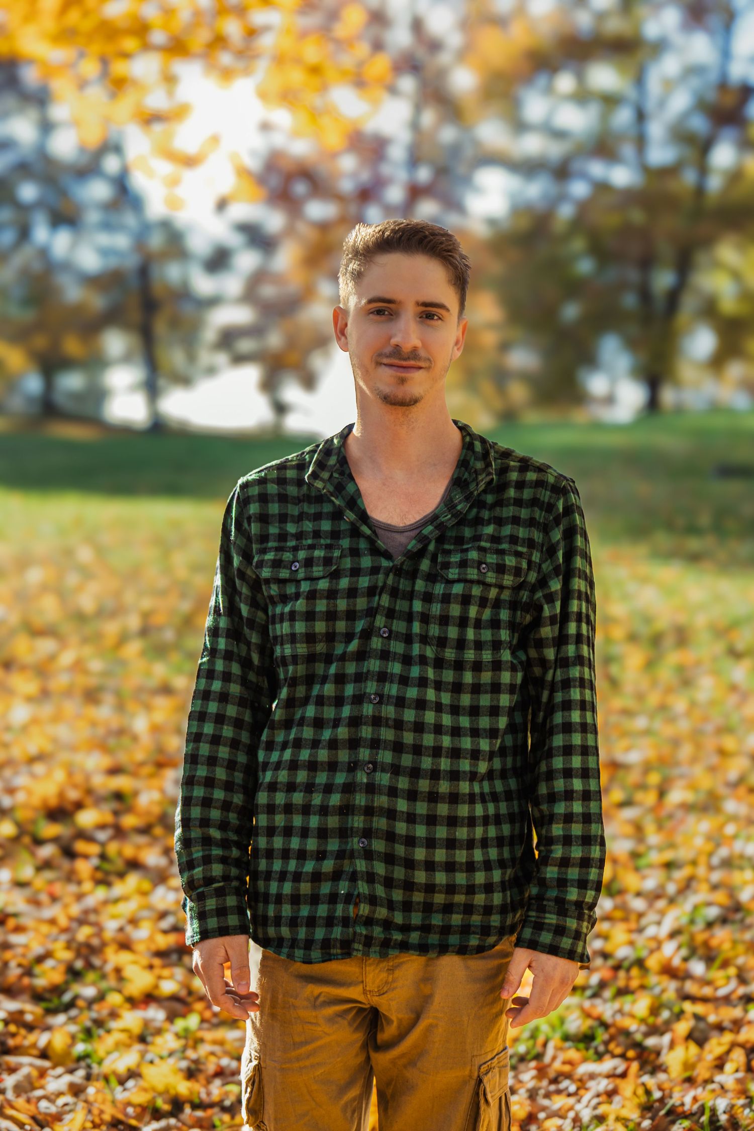 Person wearing green plaid flannel and brown pants poses in autumn leaves on sunny fall day.
