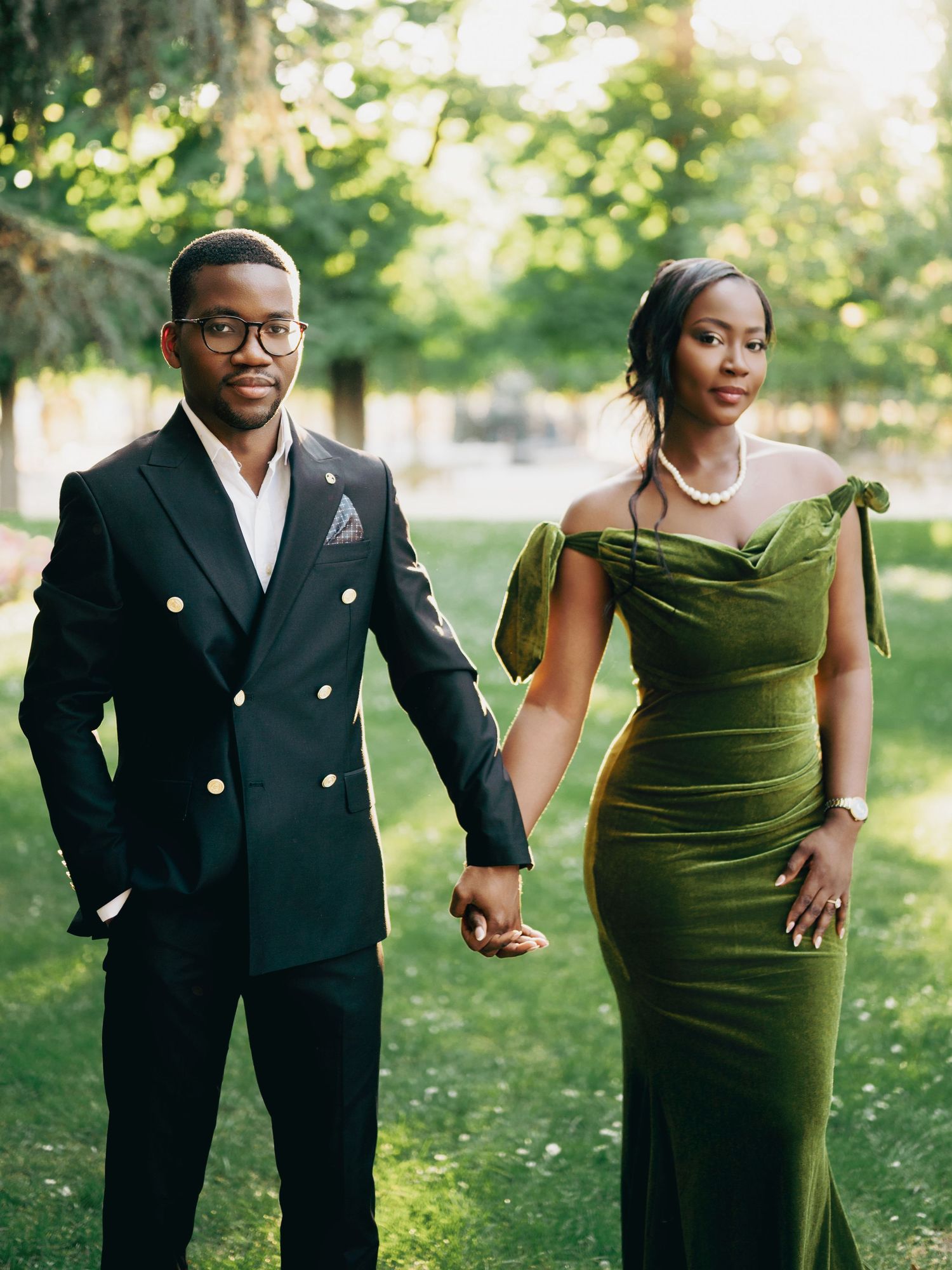 Stylish Black couple holding hands outdoors, man in black double-breasted suit, woman in olive green velvet gown.