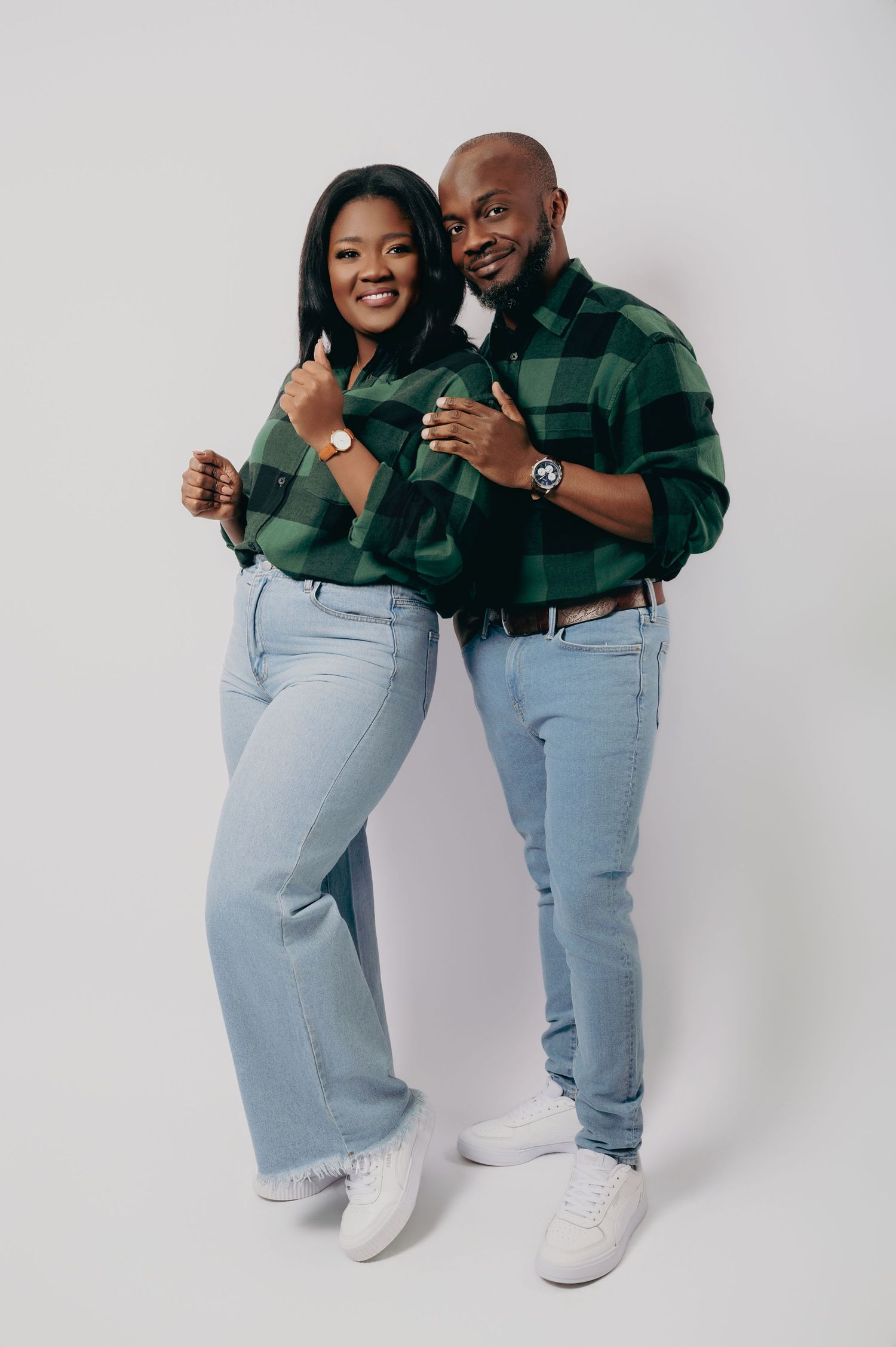 A smiling couple in matching green plaid shirts and light blue jeans posing together against a white background.