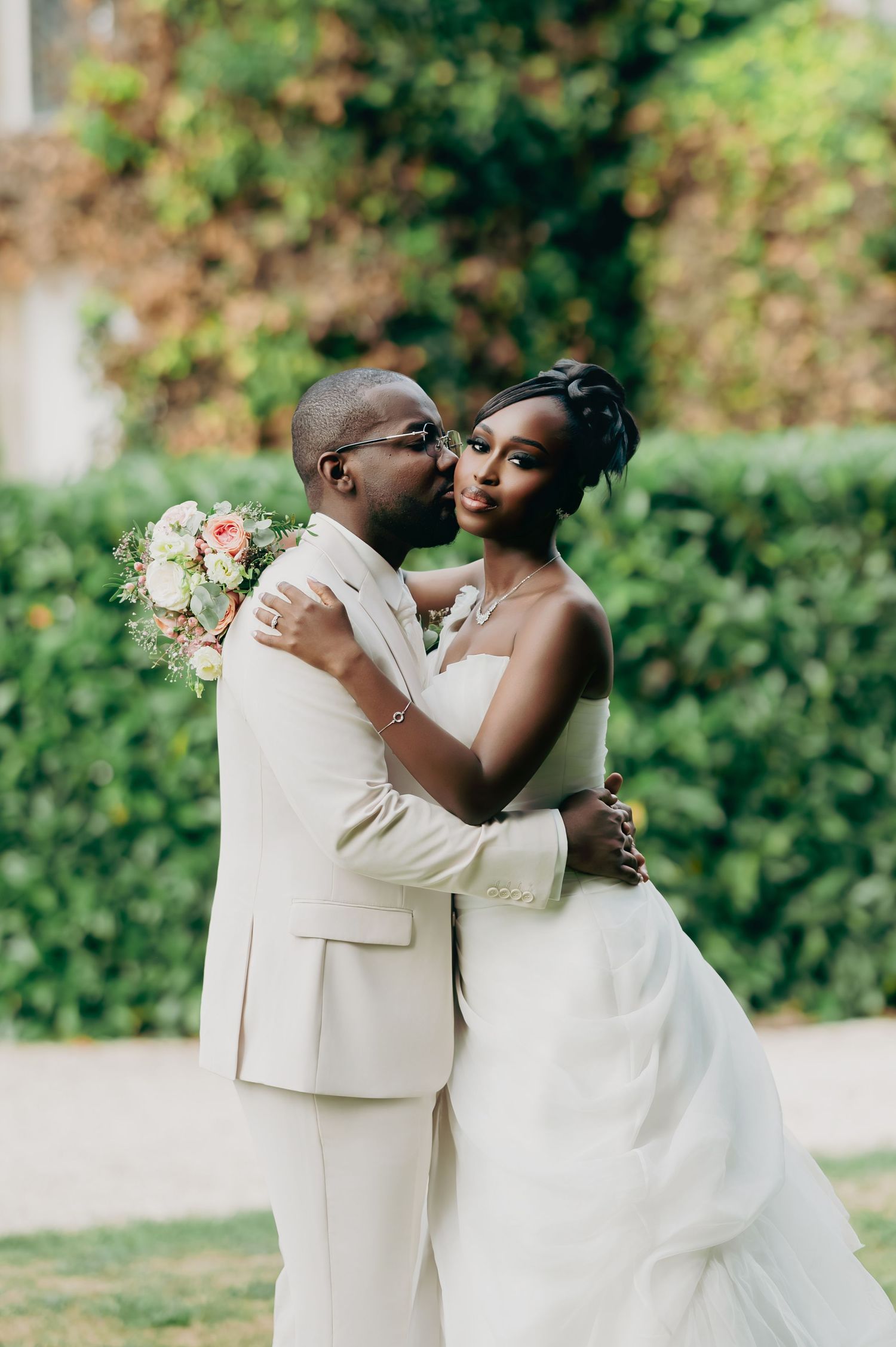 Black groom in cream suit kisses bride's cheek while holding a pink bouquet in a lush garden setting.