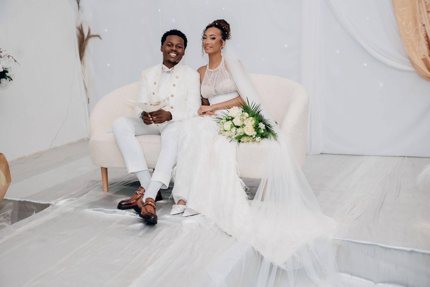 Bride in white gown with long veil and groom in white suit pose on sofa.